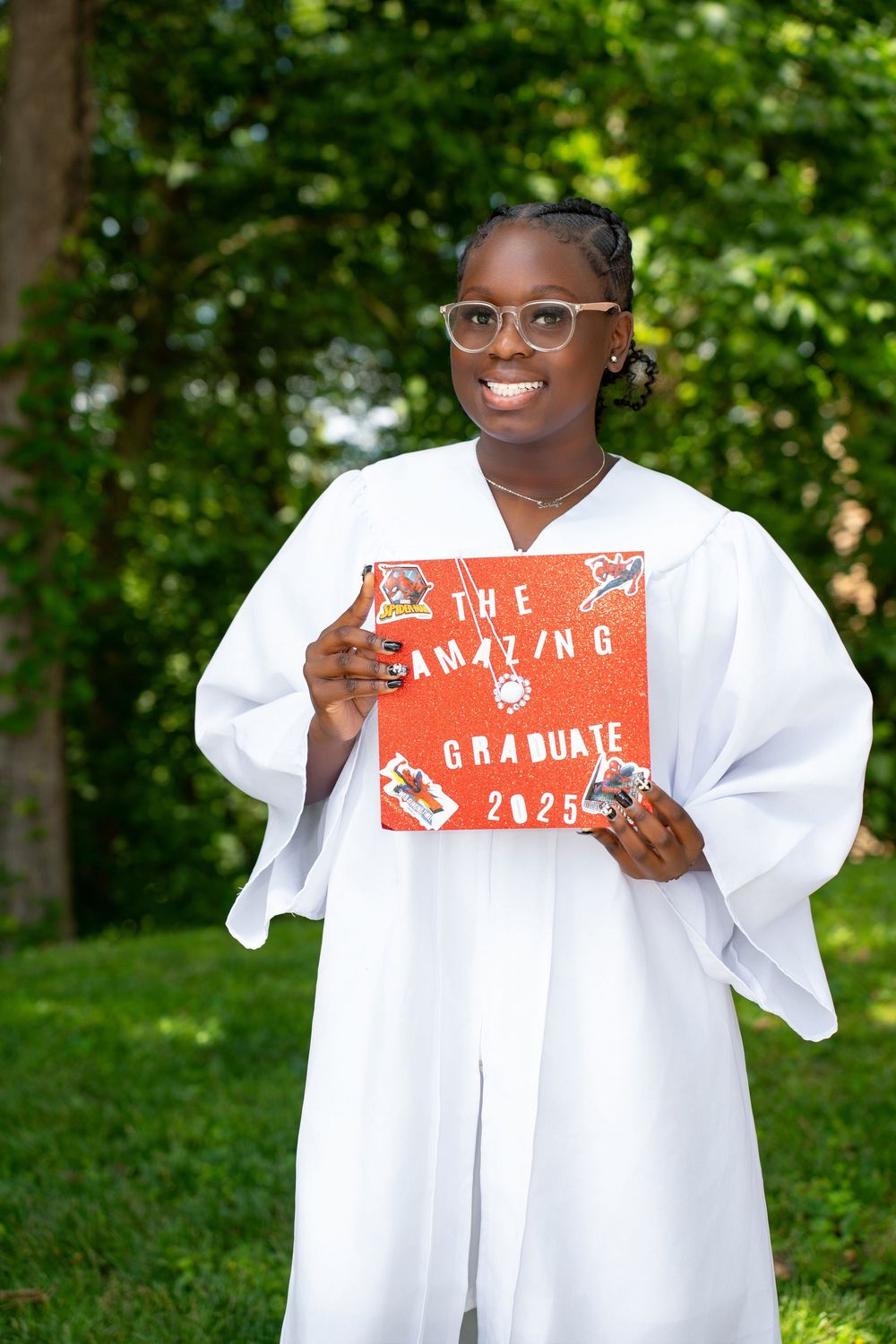 Smiling graduate in white gown holds decorated red cap reading The Amazing Graduate 2025 outdoors.