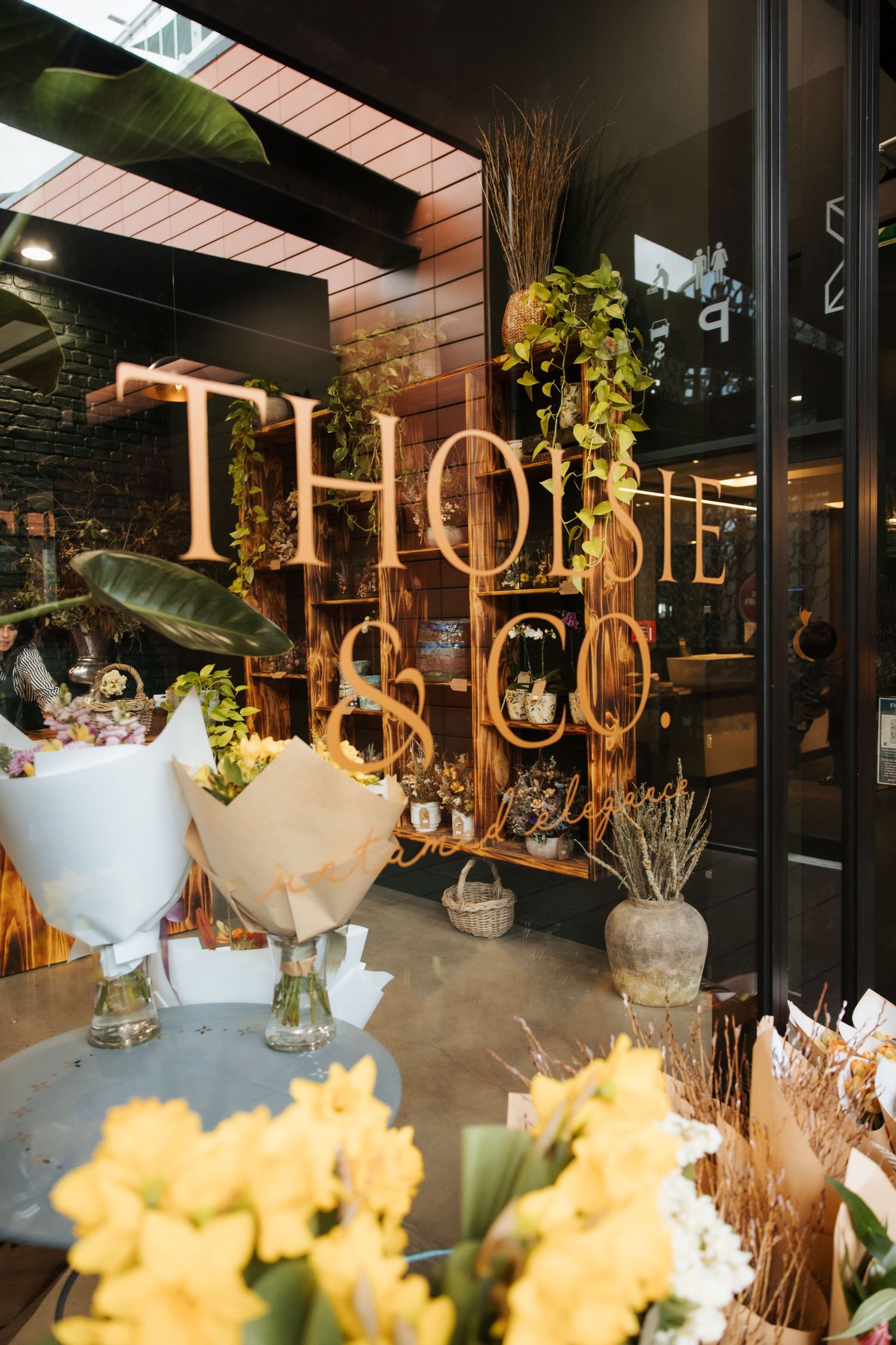 Thoisie & Co. flower shop storefront with golden signage, lush greenery, and fresh bouquets including yellow daffodils.