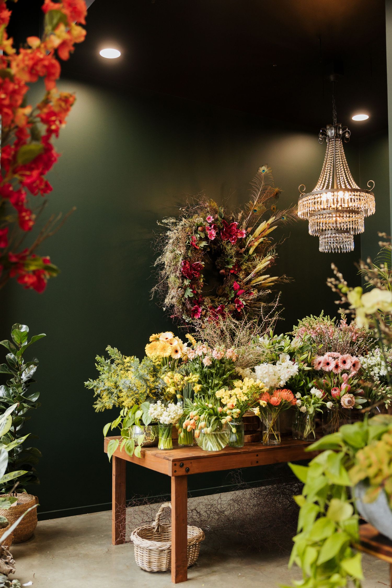 Elegant flower shop display with colorful blooms on wooden table, crystal chandelier, and floral wreath against dark green wall.