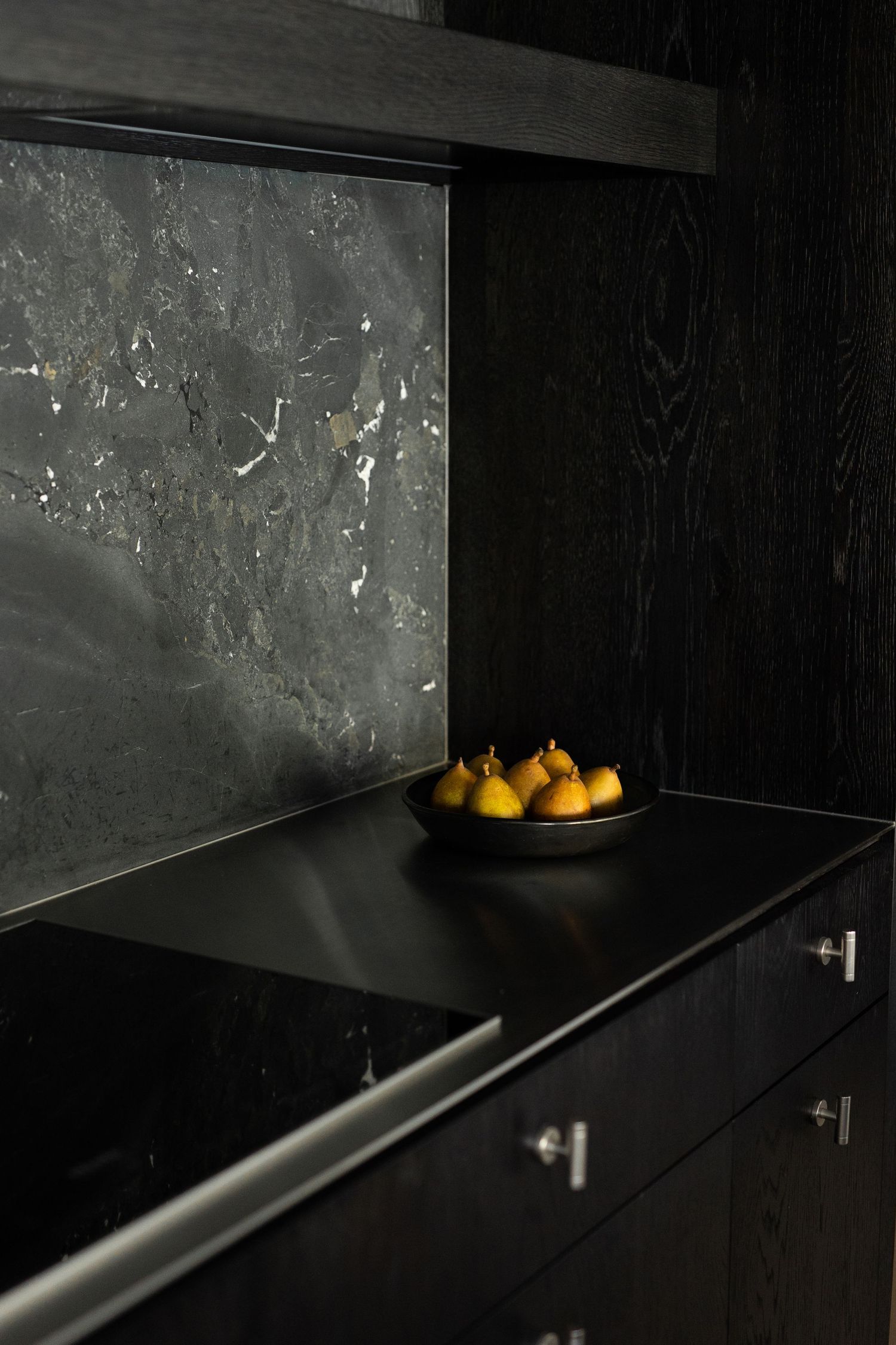 Dark luxury kitchen with black marble backsplash, ebony wood cabinets, and a bowl of yellow pears on granite countertop.