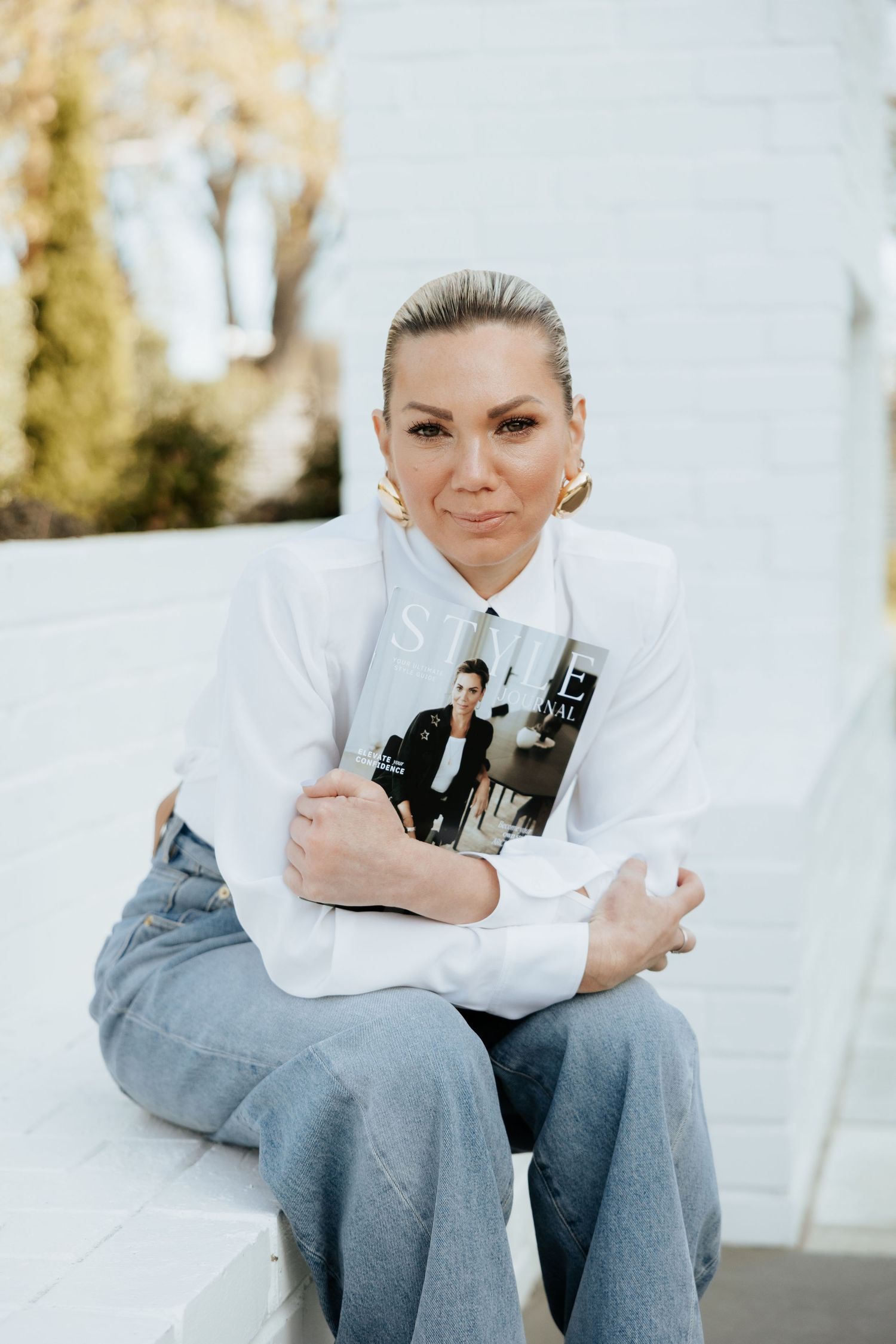 Woman in white shirt and jeans sits on steps holding a Style magazine, wearing gold hoop earrings.