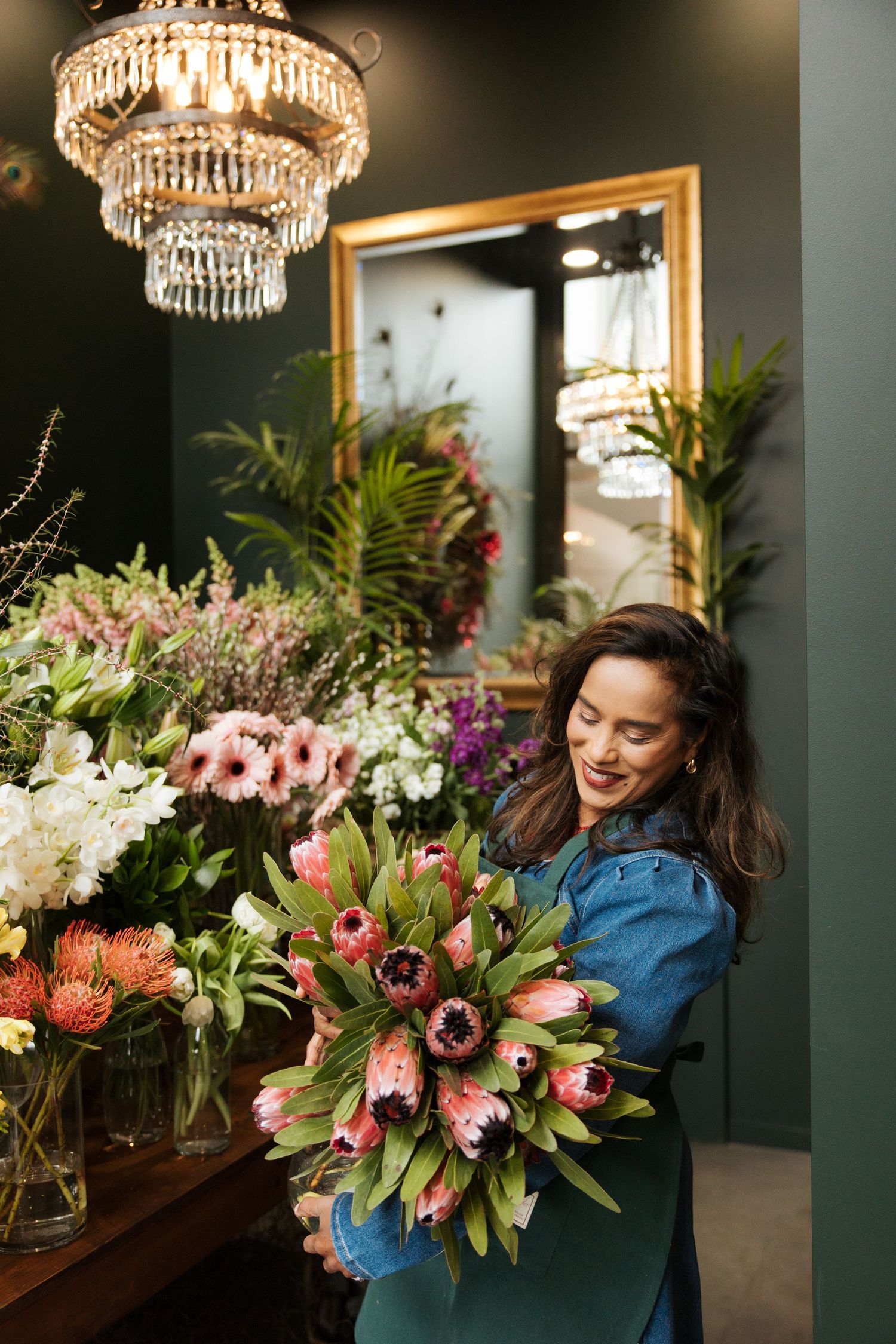 Florist in denim apron arranging pink tulip bouquet in elegant flower shop with chandelier and mirror.