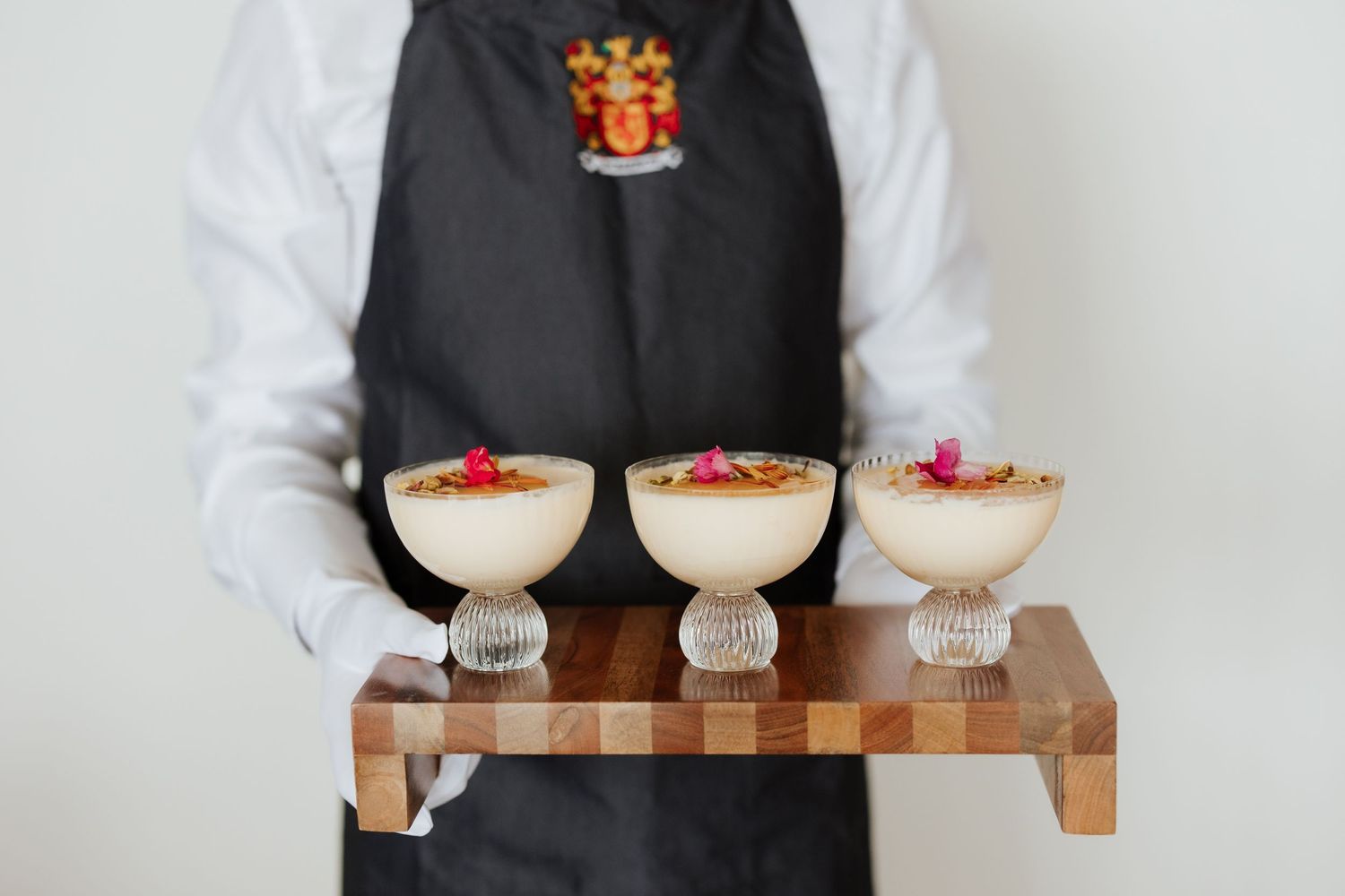 Server in black apron holding wooden tray with three elegant coupe glasses topped with floral garnishes.