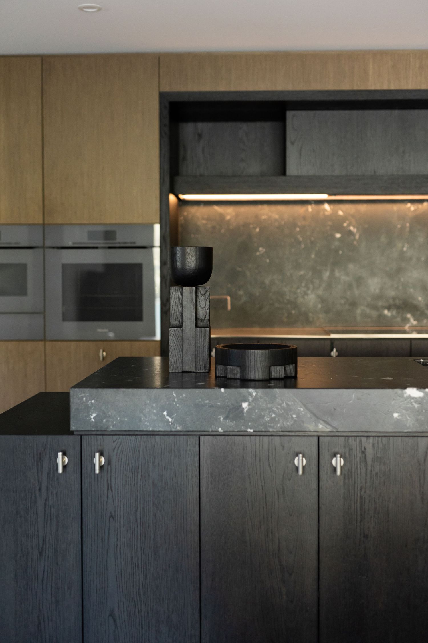 Modern kitchen with dark oak cabinets, marble island, stainless steel ovens, and warm under-cabinet lighting.