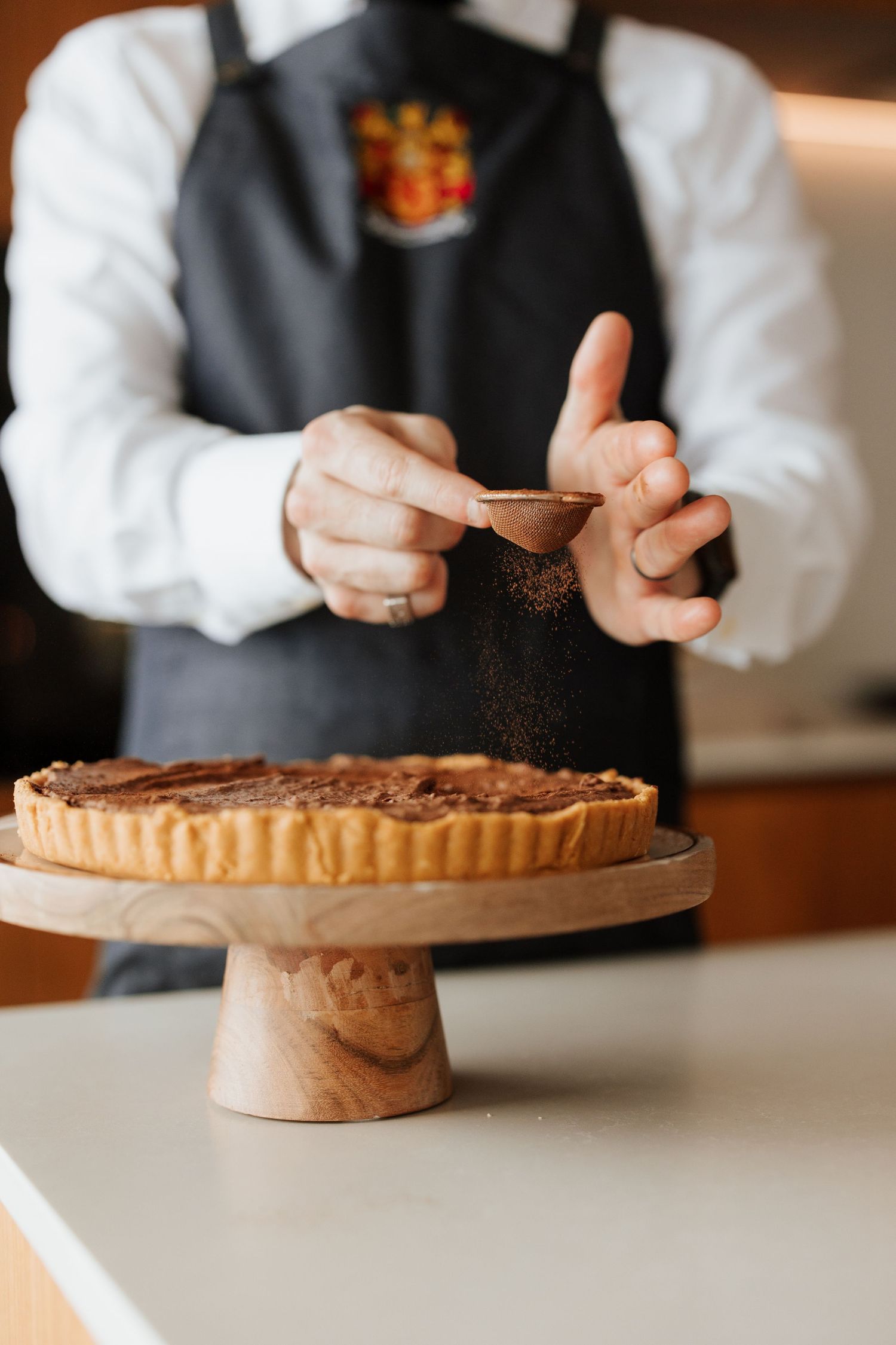 Chef dusting cocoa powder over a chocolate tart on a wooden cake stand in a professional kitchen.