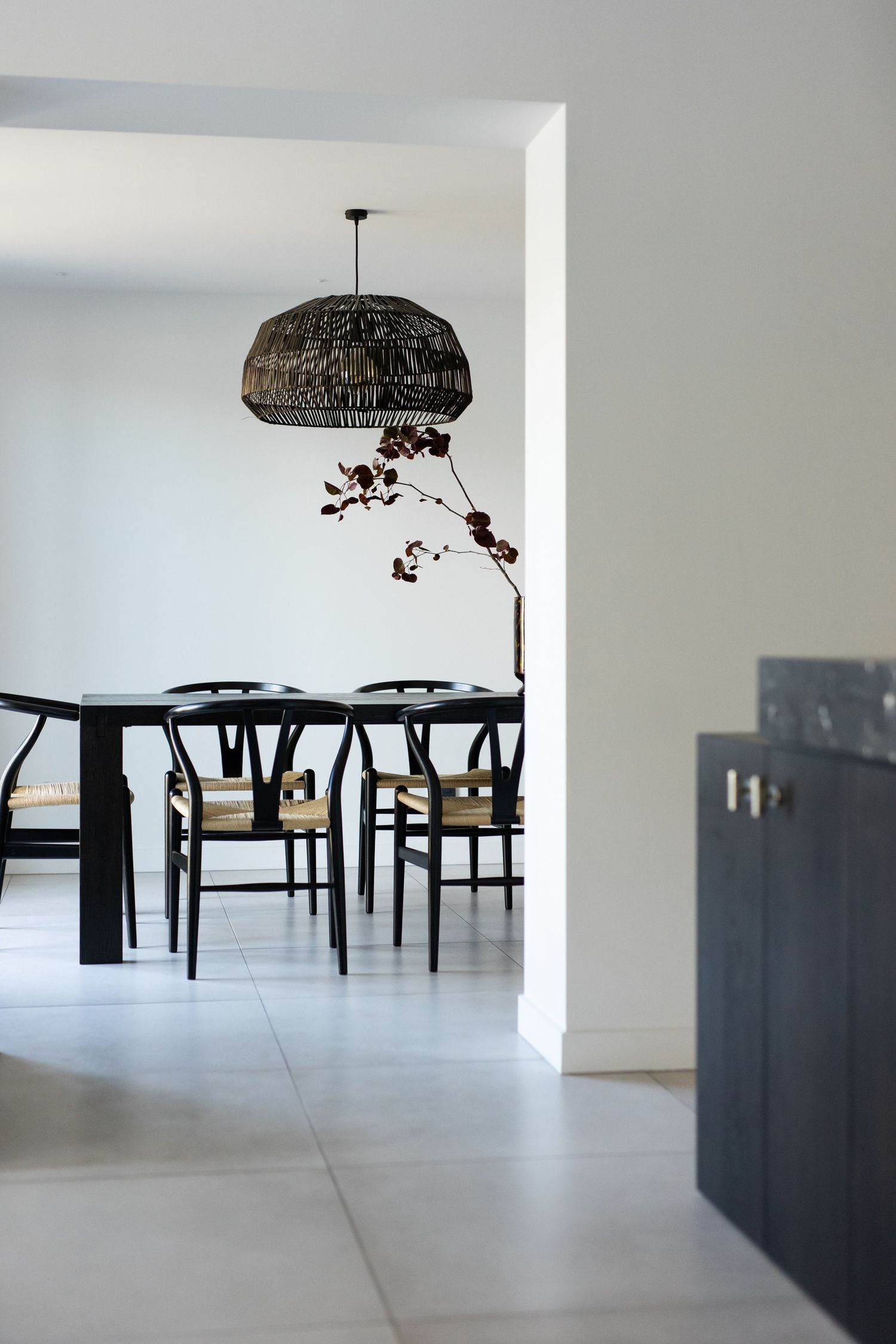Modern dining room with black table, Wishbone chairs, woven pendant light, and a decorative branch centerpiece.