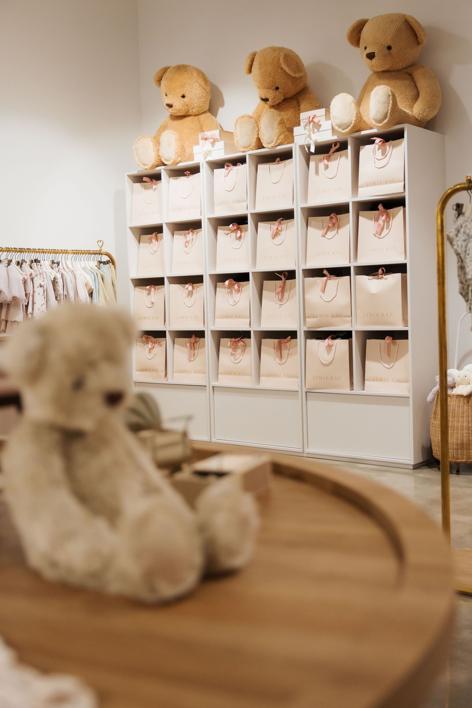 Cozy baby boutique with teddy bears on white cubby shelves, fabric storage bins, and soft clothing rack in warm neutral tones.