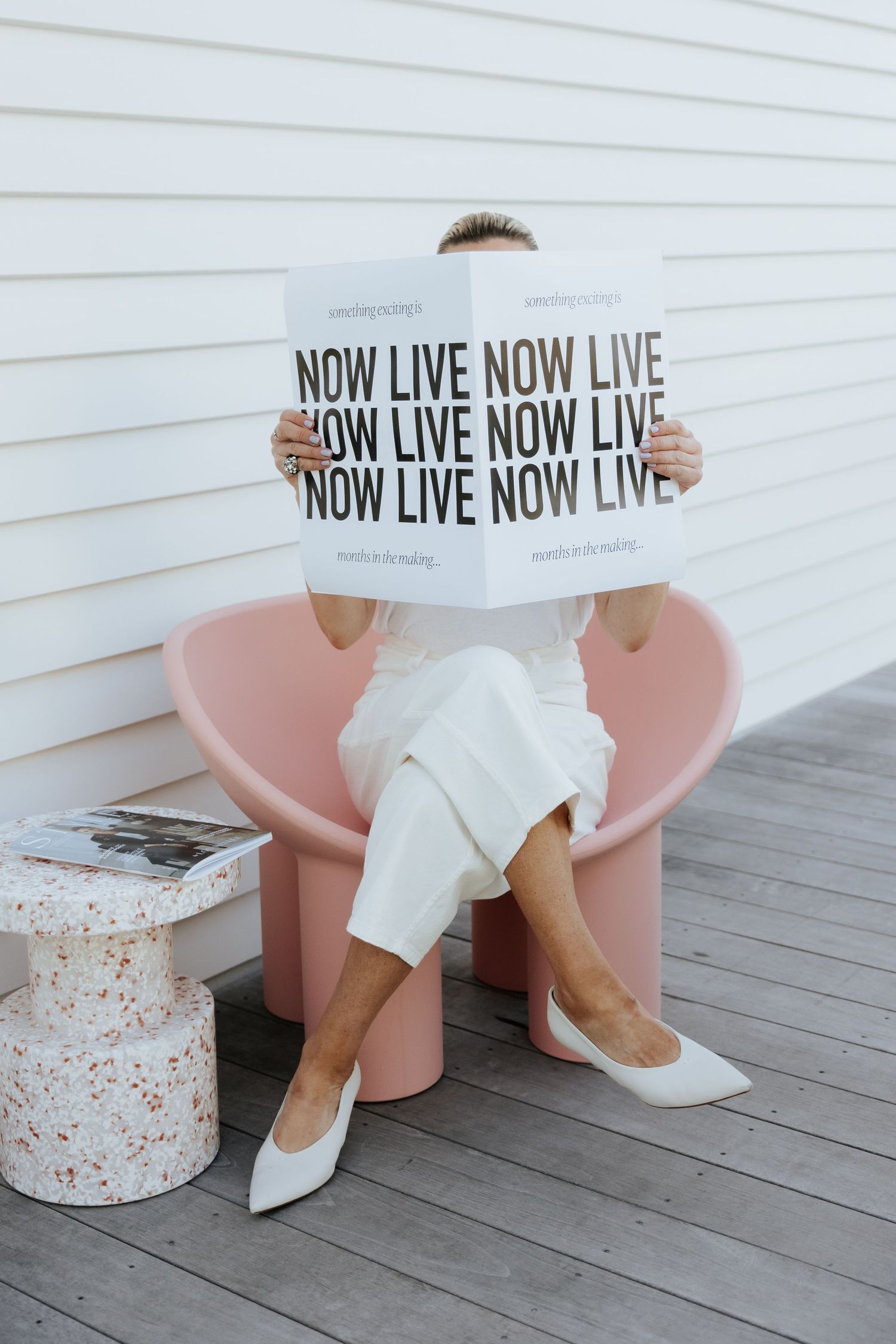 Person in white outfit sitting in pink chair holding a 'Now Live' sign outdoors on a wooden deck.