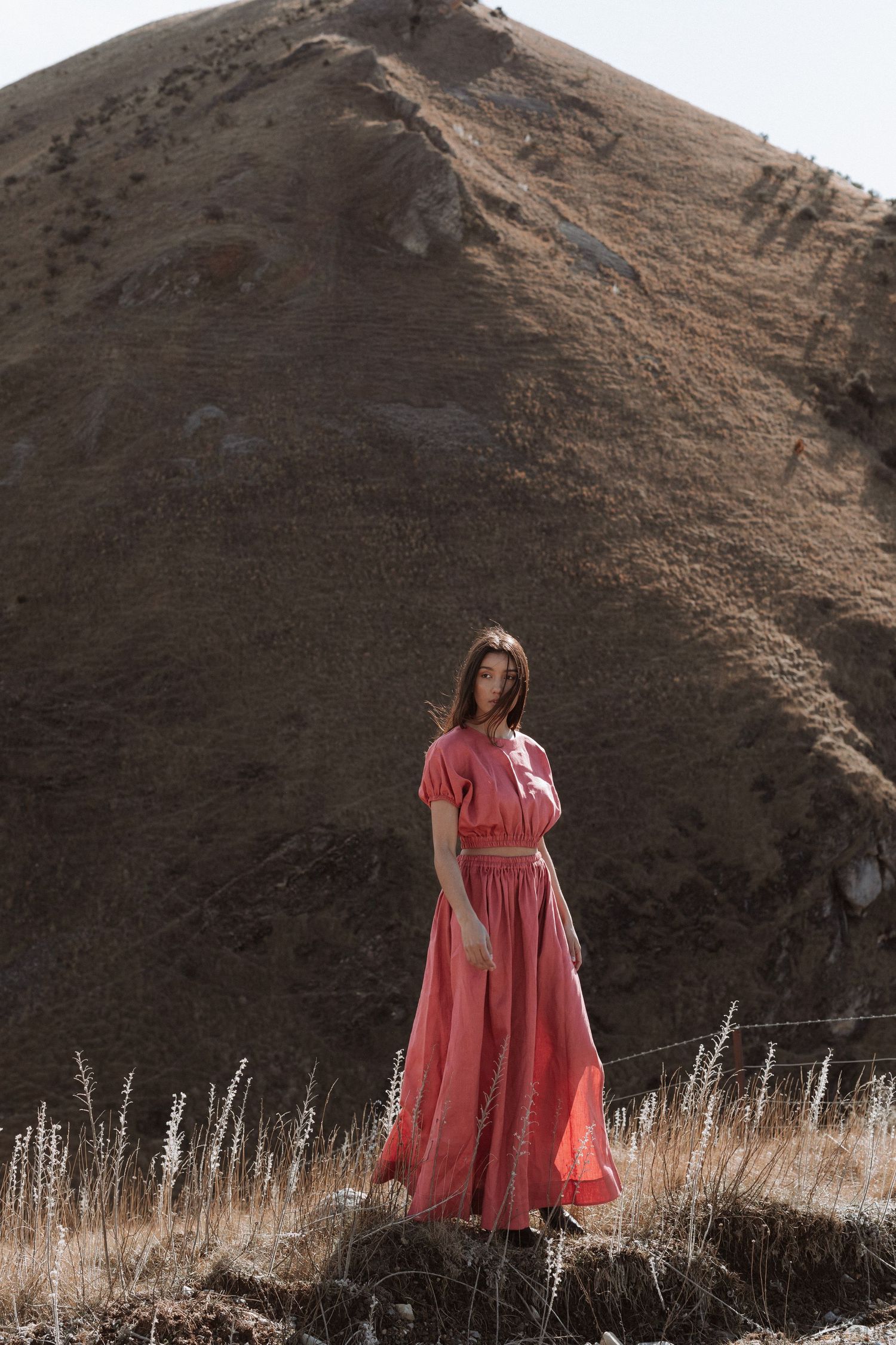 Woman in flowing pink maxi dress standing on rocky hillside terrain in natural outdoor landscape setting.