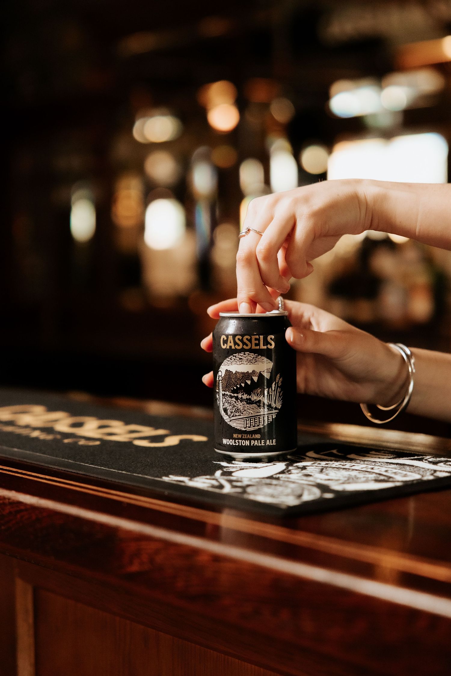 Hands opening a Cassels Woolston Pale Ale can on a wooden bar counter in a warmly lit pub.