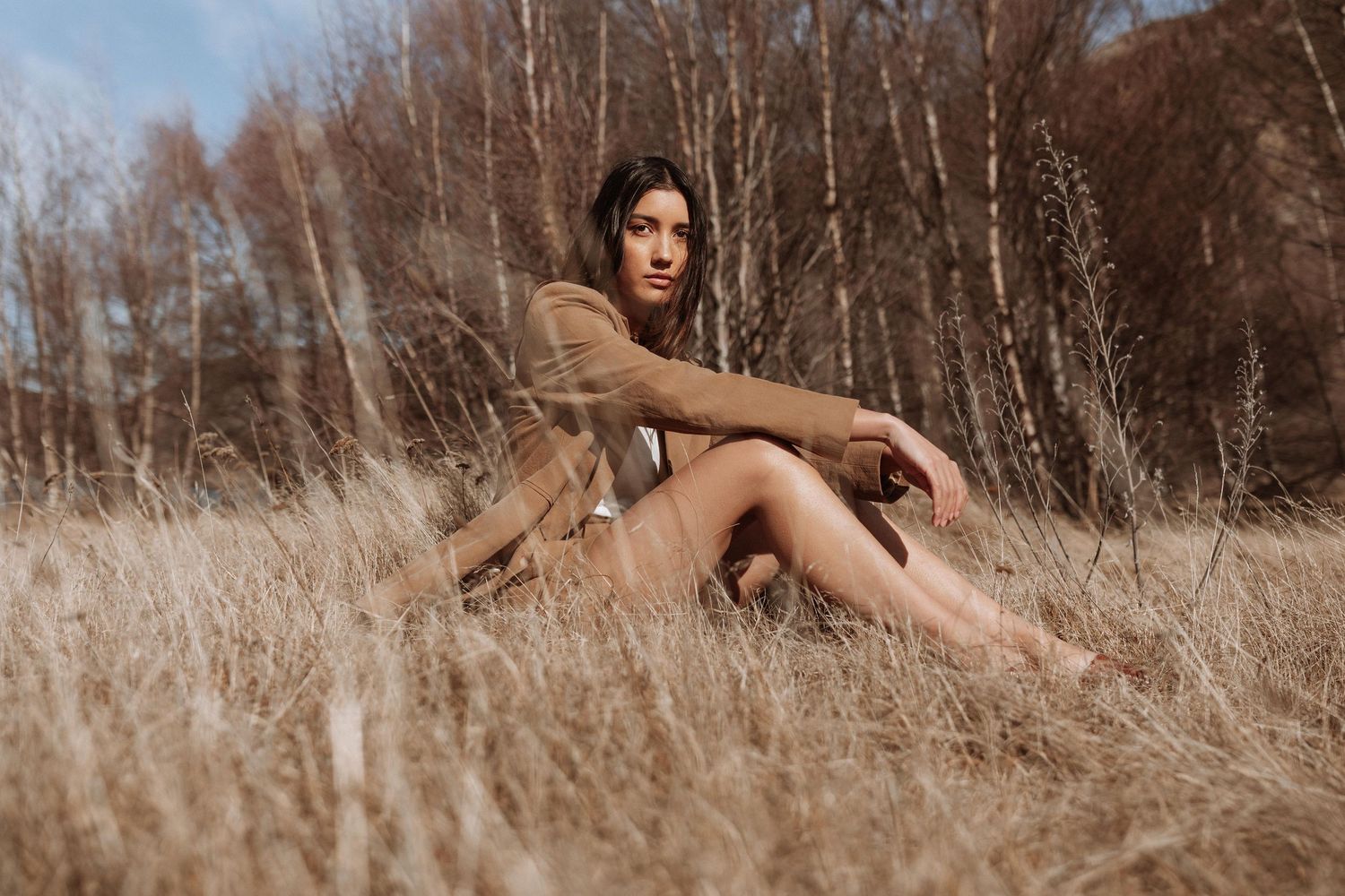 A young woman in a brown coat sits barefoot in golden dry grass with bare trees in the background.