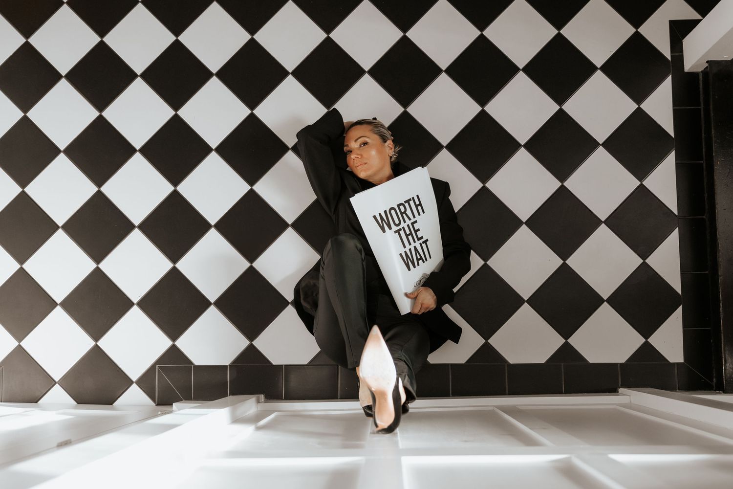 Person in black suit holds 'Worth The Wait' sign against bold black and white checkered wall pattern.