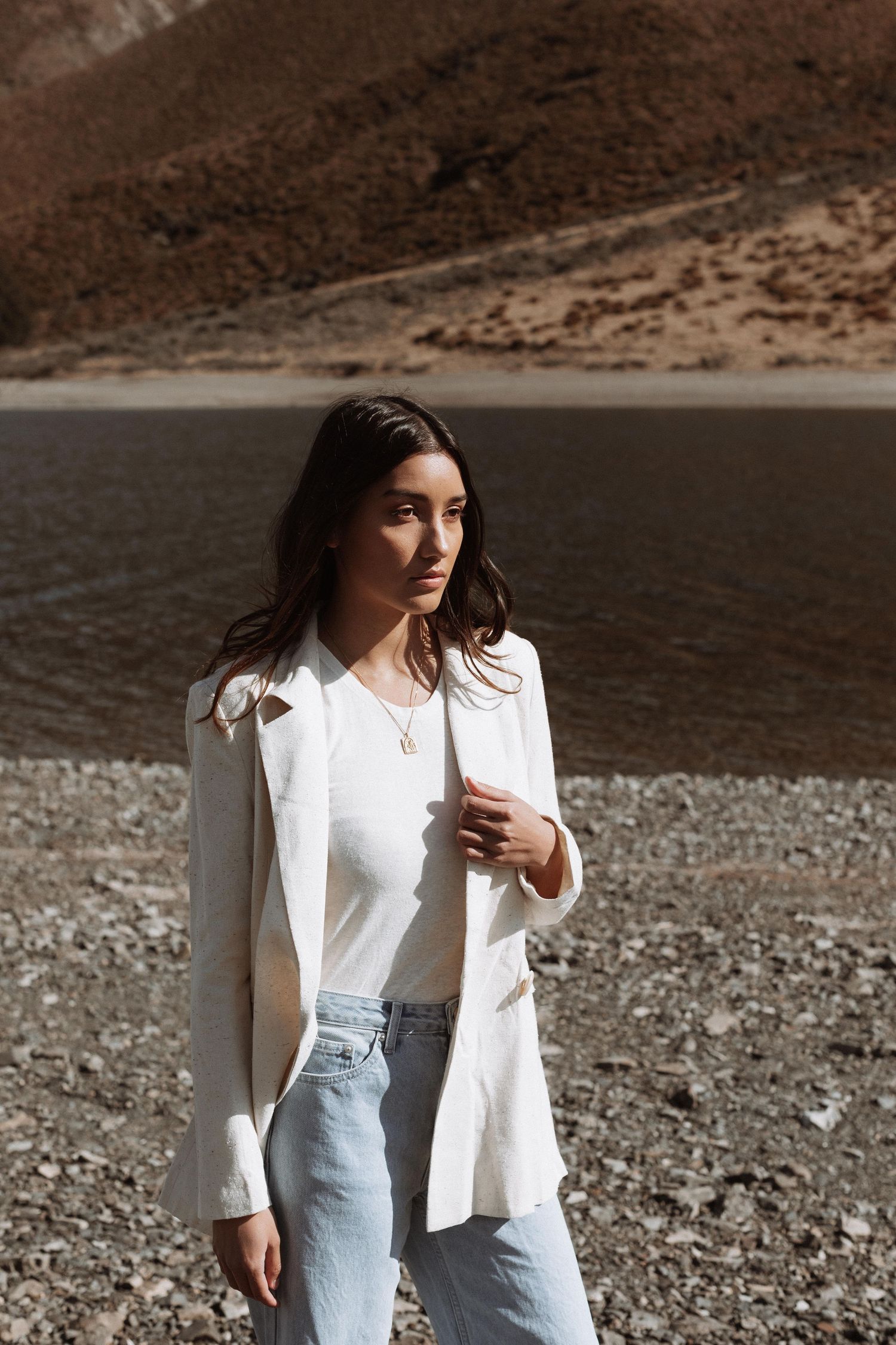 Woman in white blazer and jeans standing on rocky shore with mountain lake backdrop in sunlight.