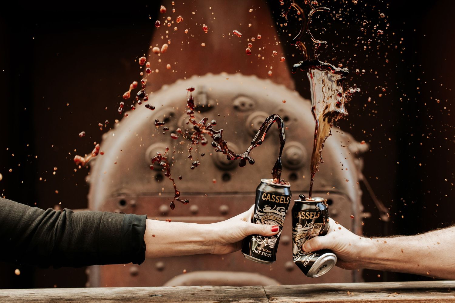 Two hands toasting Castle Black beer cans with dramatic liquid splash against rustic copper background.