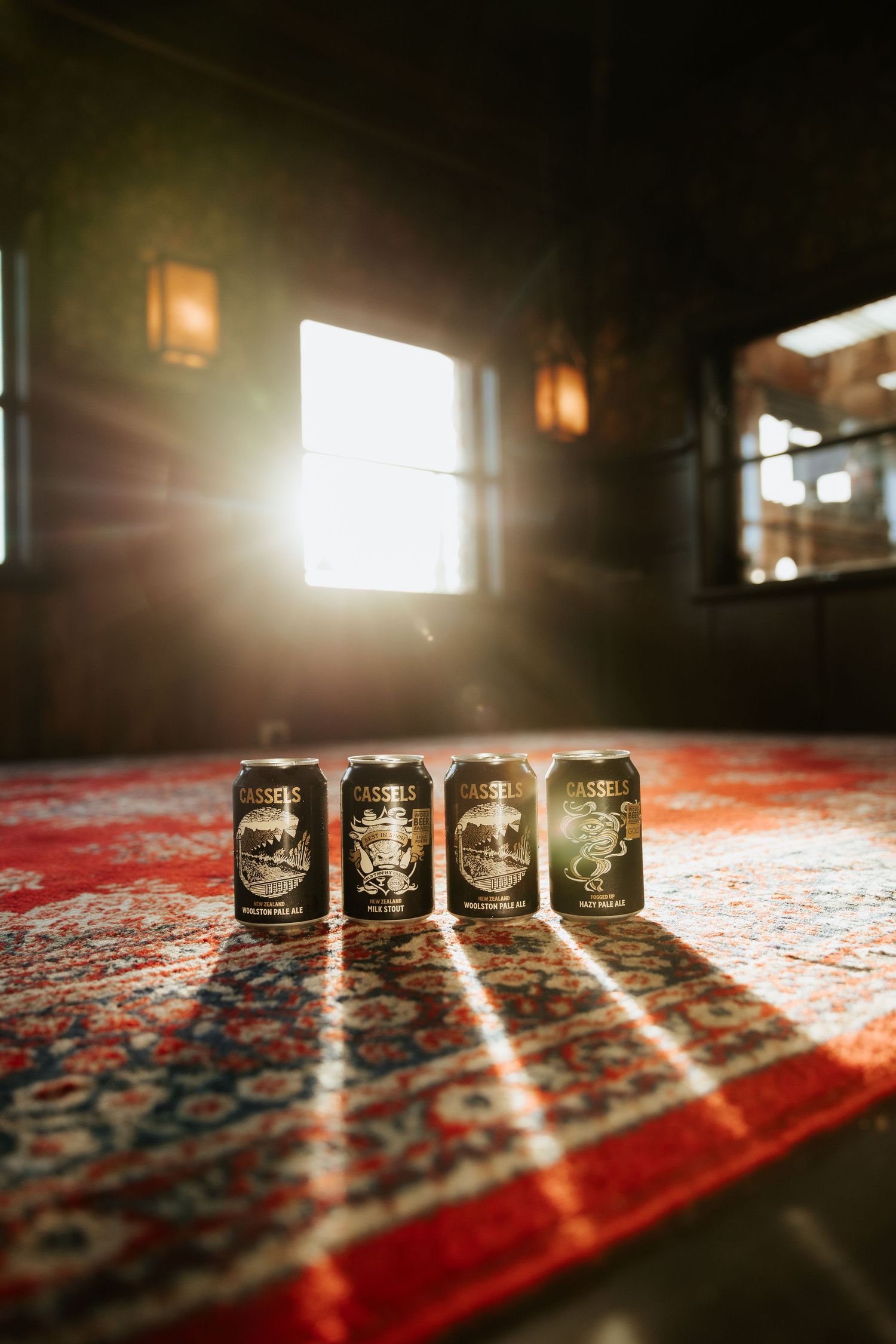 Four Cassels beer cans arranged on a red patterned rug with dramatic sunlight streaming through windows in a dark room.