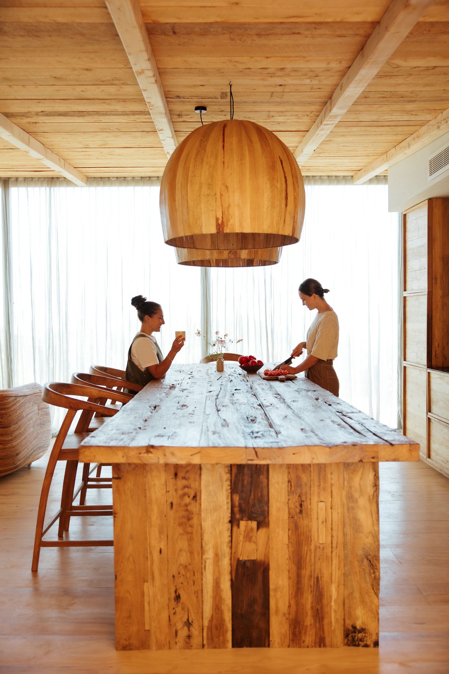 Two women dining at a rustic wooden table beneath a large dome pendant light in a modern wood-paneled room.
