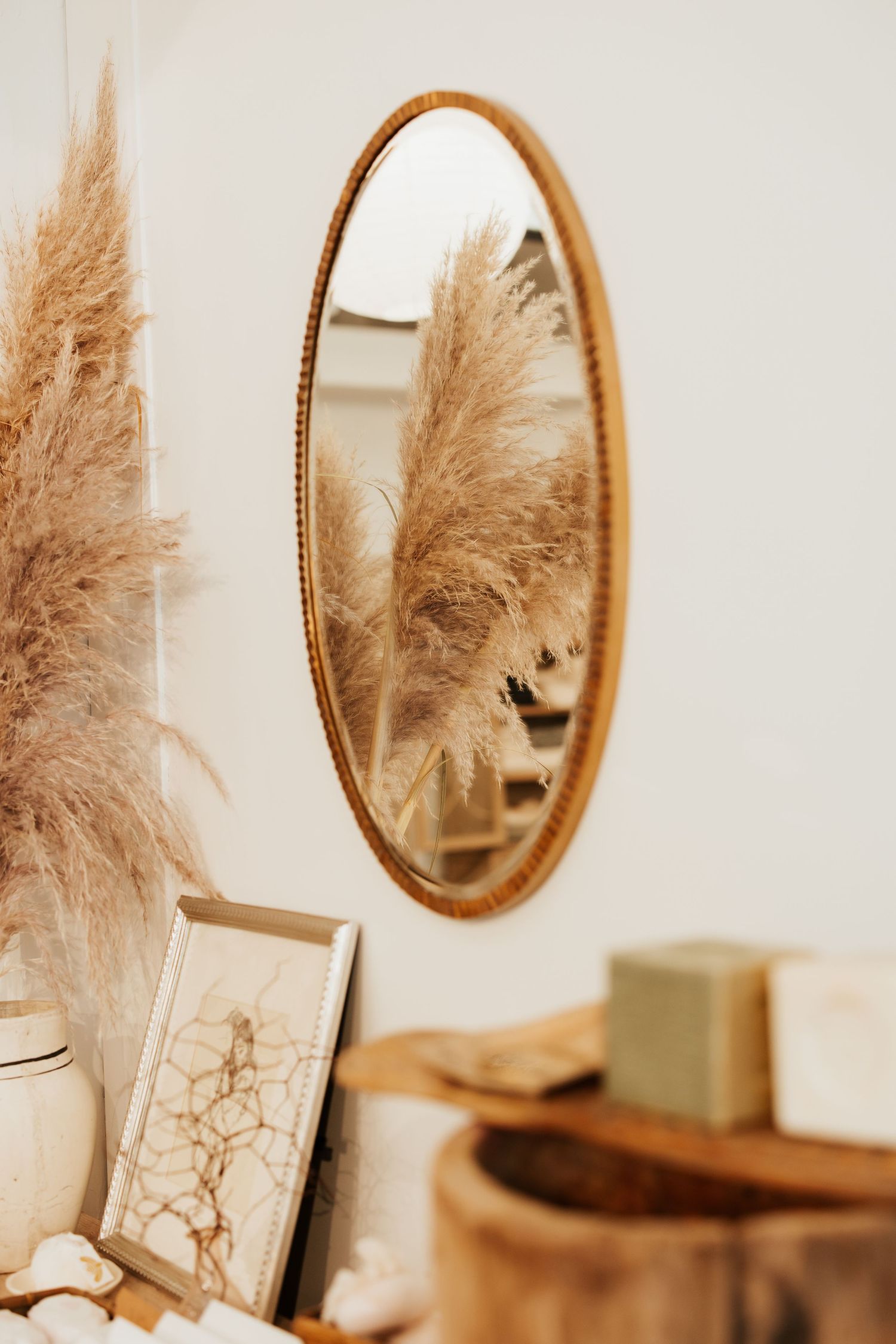 Oval gold-framed mirror reflecting pampas grass, displayed above a wooden shelf with decorative items.