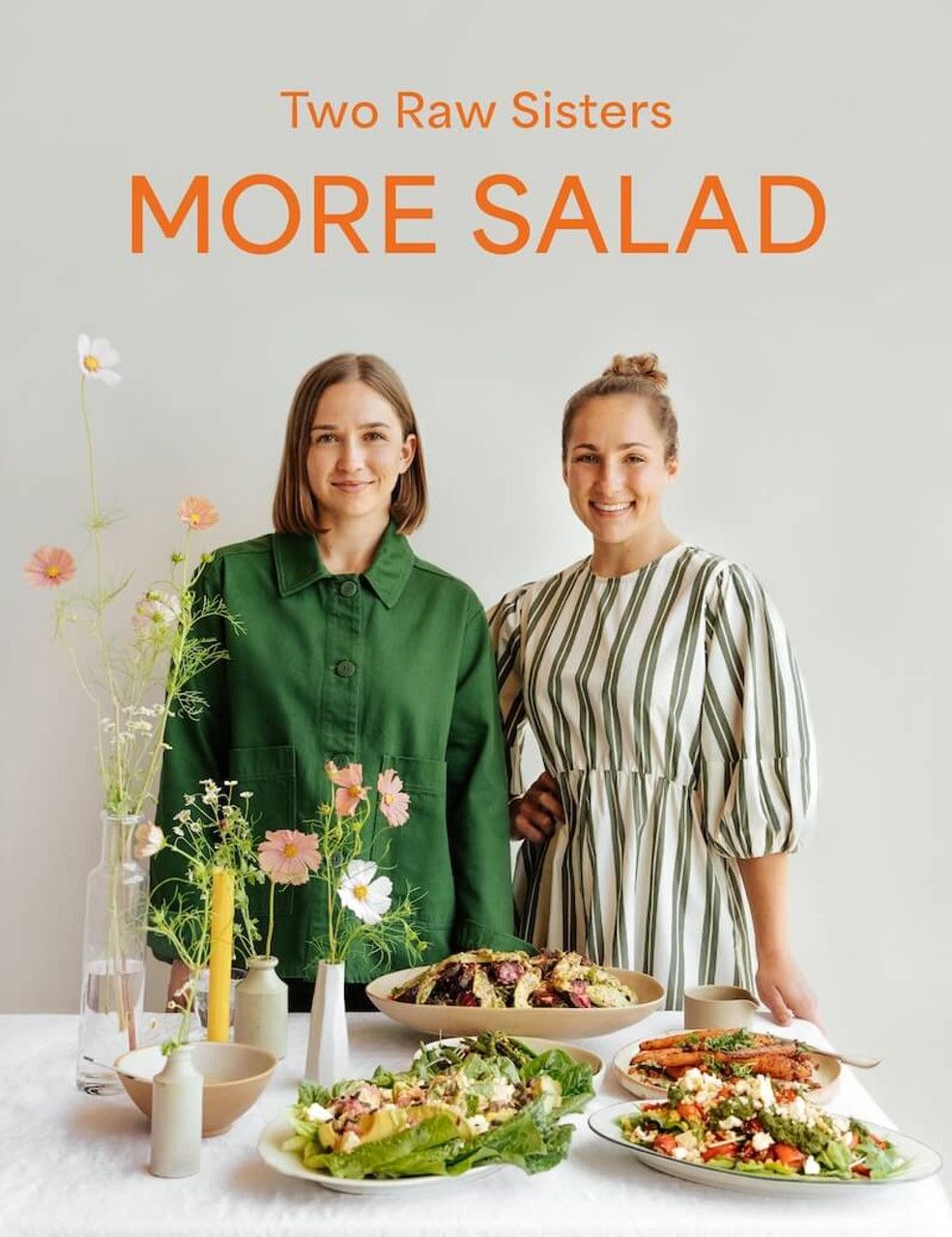 Two Raw Sisters cookbook More Salad cover featuring two women posing with colorful salad dishes on a white table.