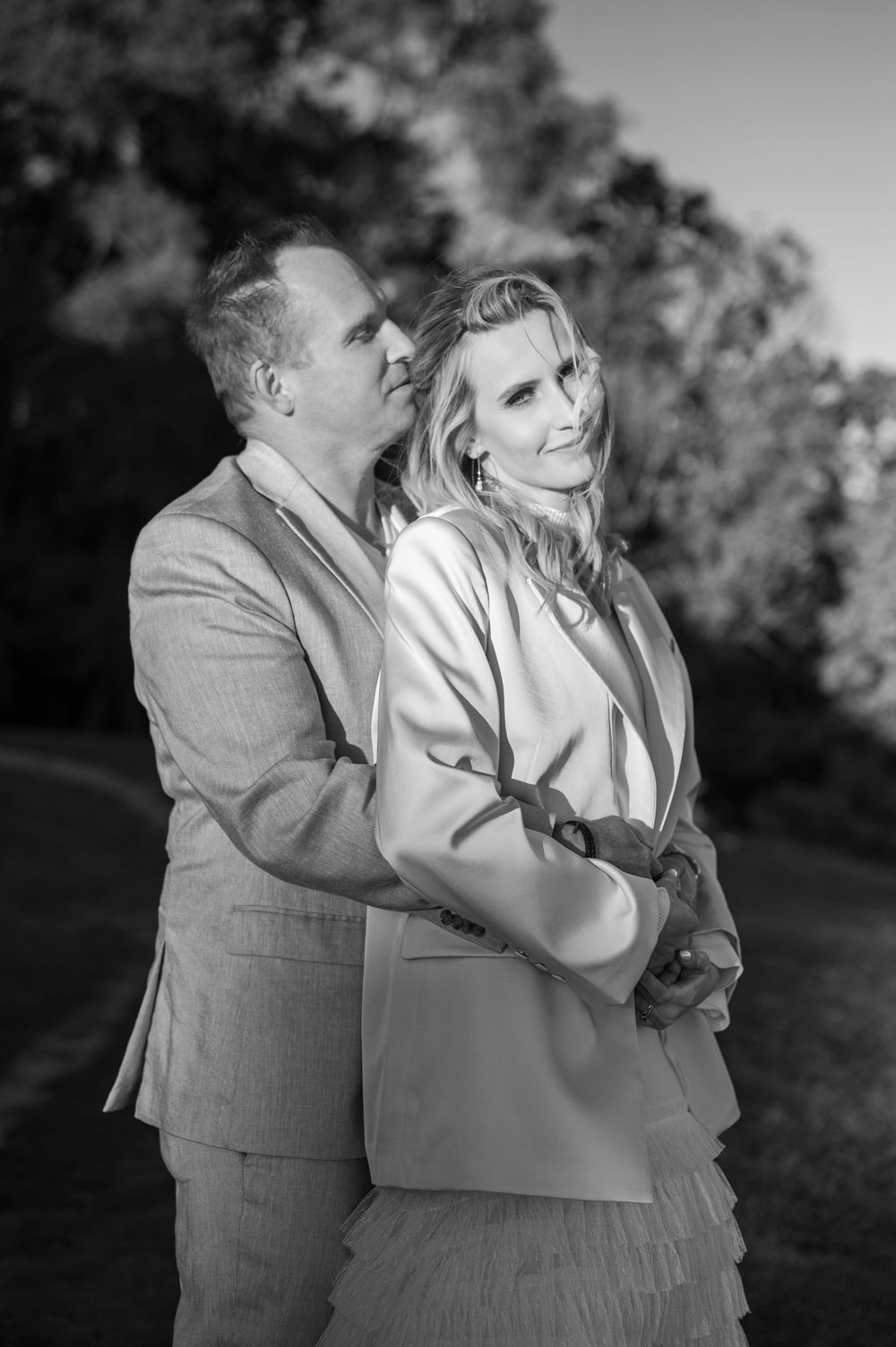 Black and white photo of a couple embracing outdoors, man in suit hugging woman in blazer, trees in background.