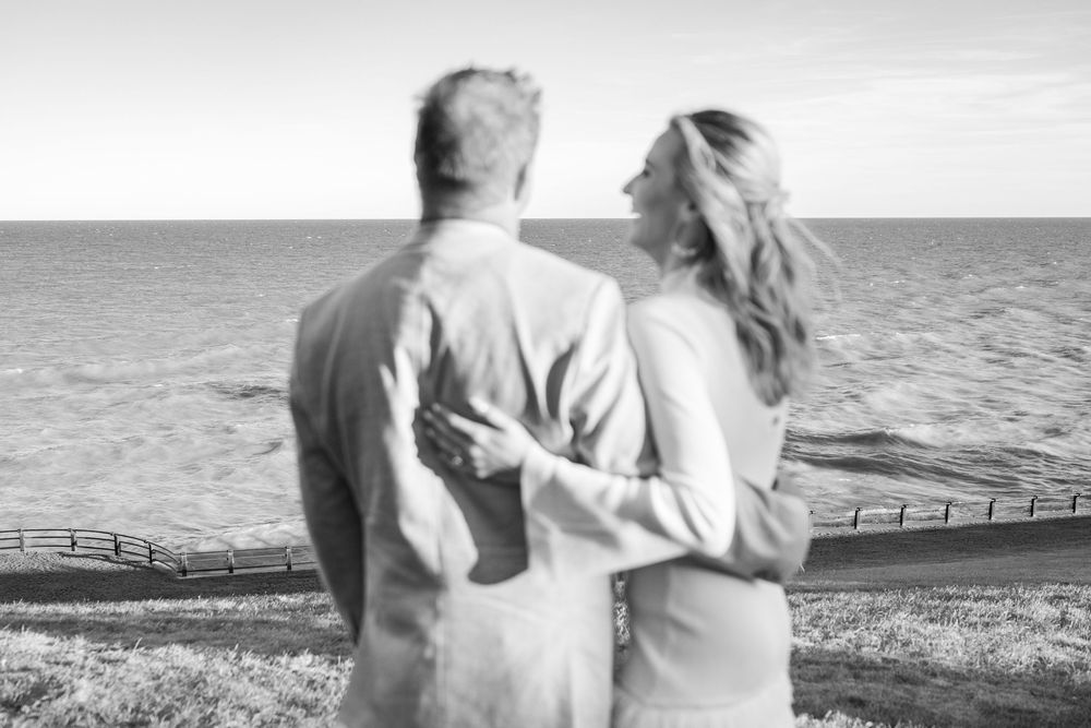 Black and white photo of a couple embracing while gazing at the ocean from a coastal overlook.