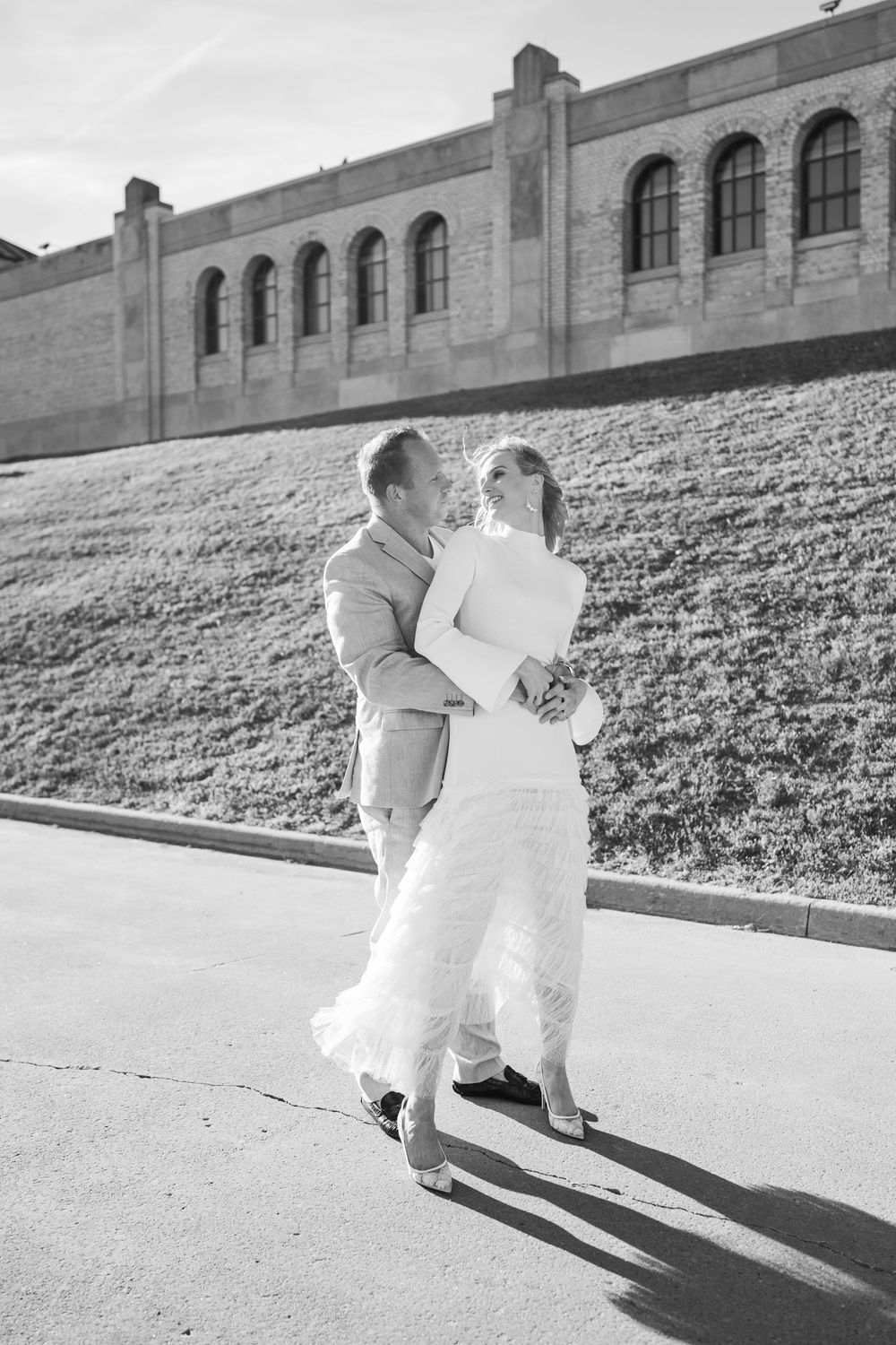 Black and white photo of a couple in wedding attire embracing outdoors near a historic stone building on a sunny day.