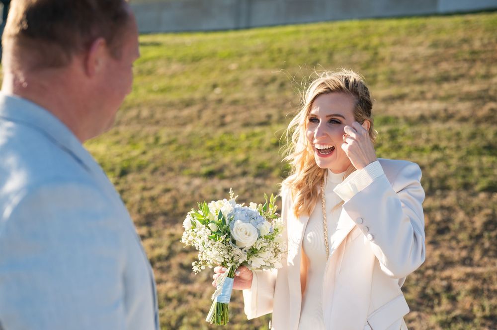 Emotional bride wiping tears while smiling, holding white floral bouquet outdoors on wedding day.