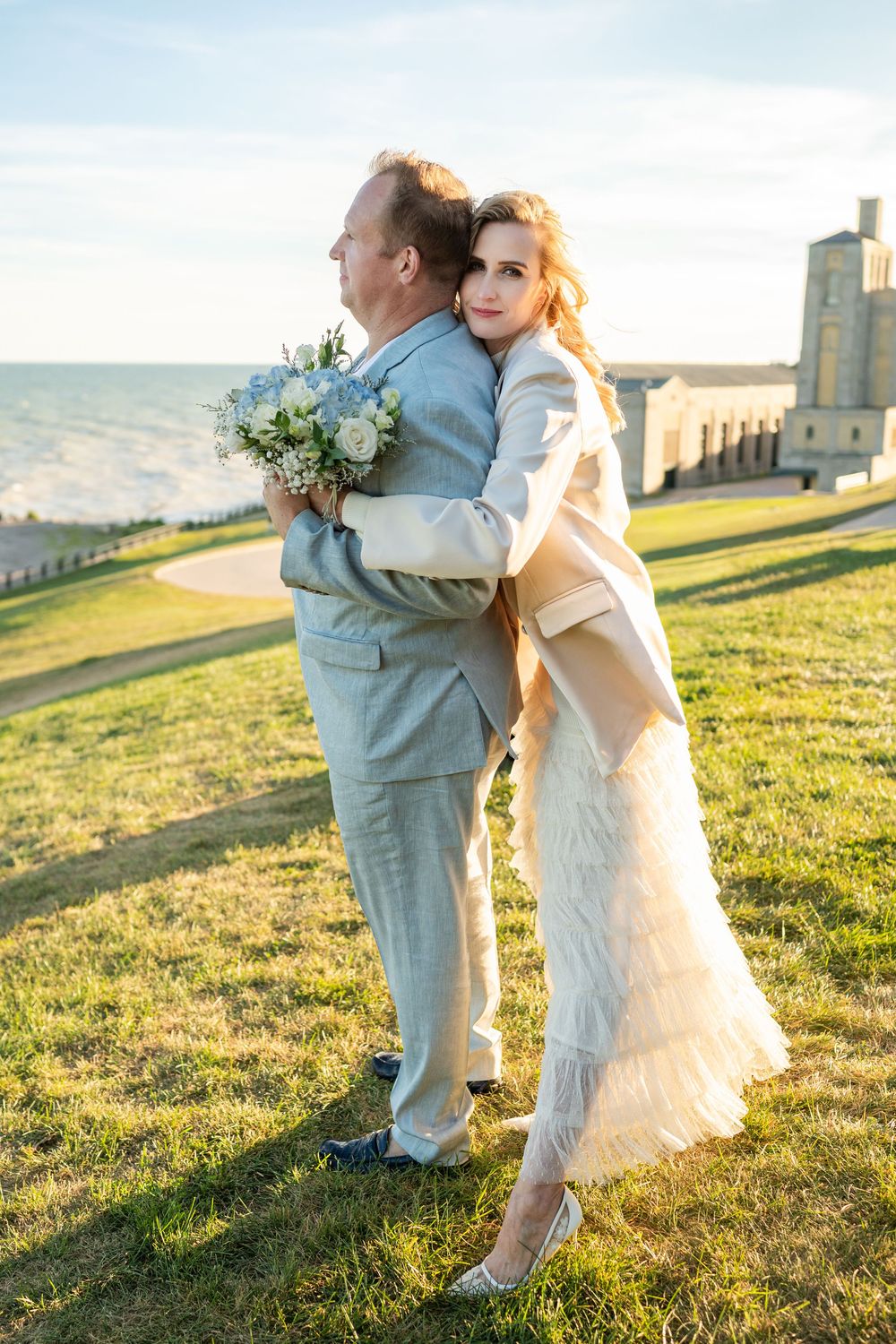 Bride in white dress hugs groom in gray suit outdoors, holding bouquet, with green lawn and historic building backdrop.