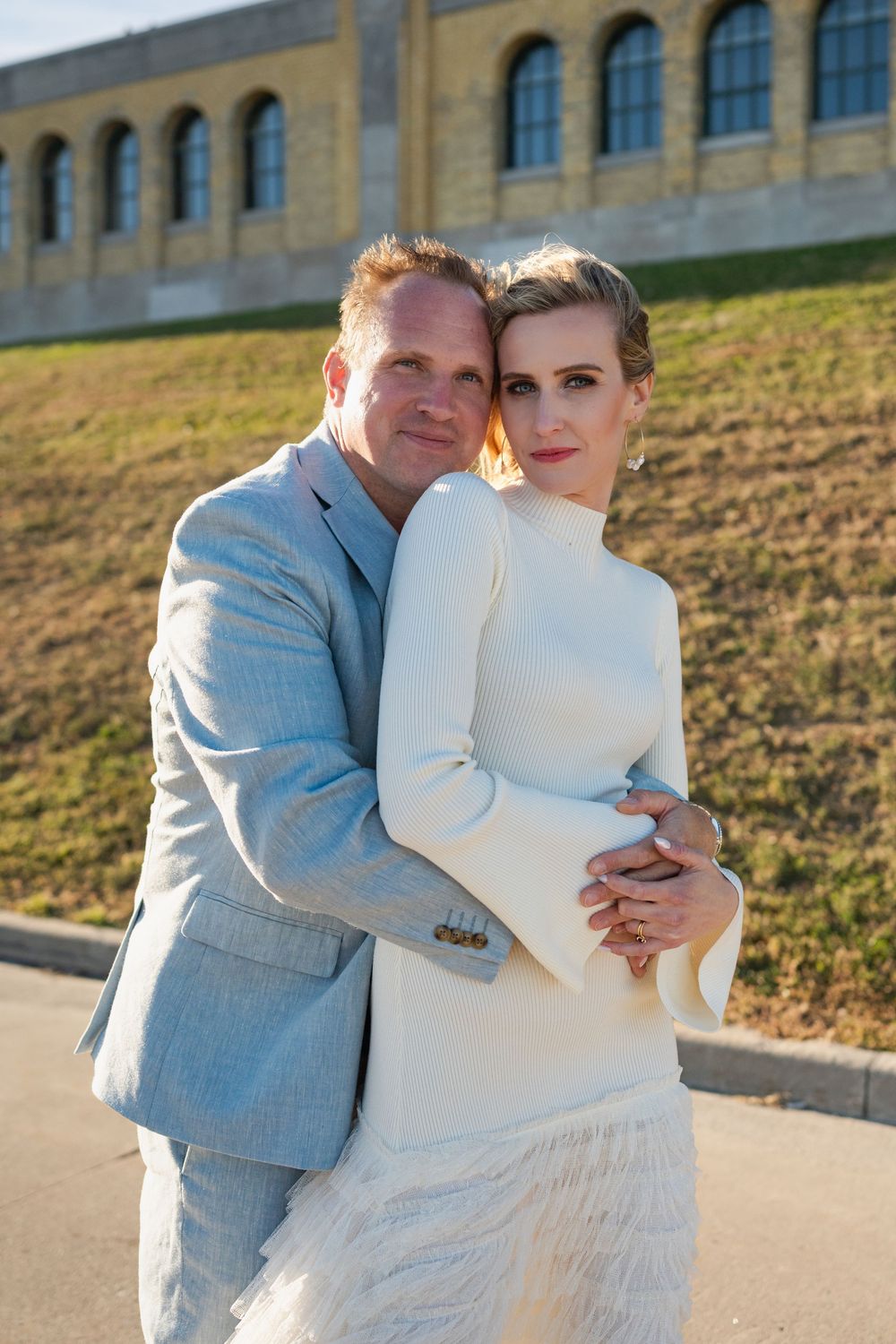 Elegant couple embracing outdoors, man in light gray suit hugging woman in white dress, posing near historic building.