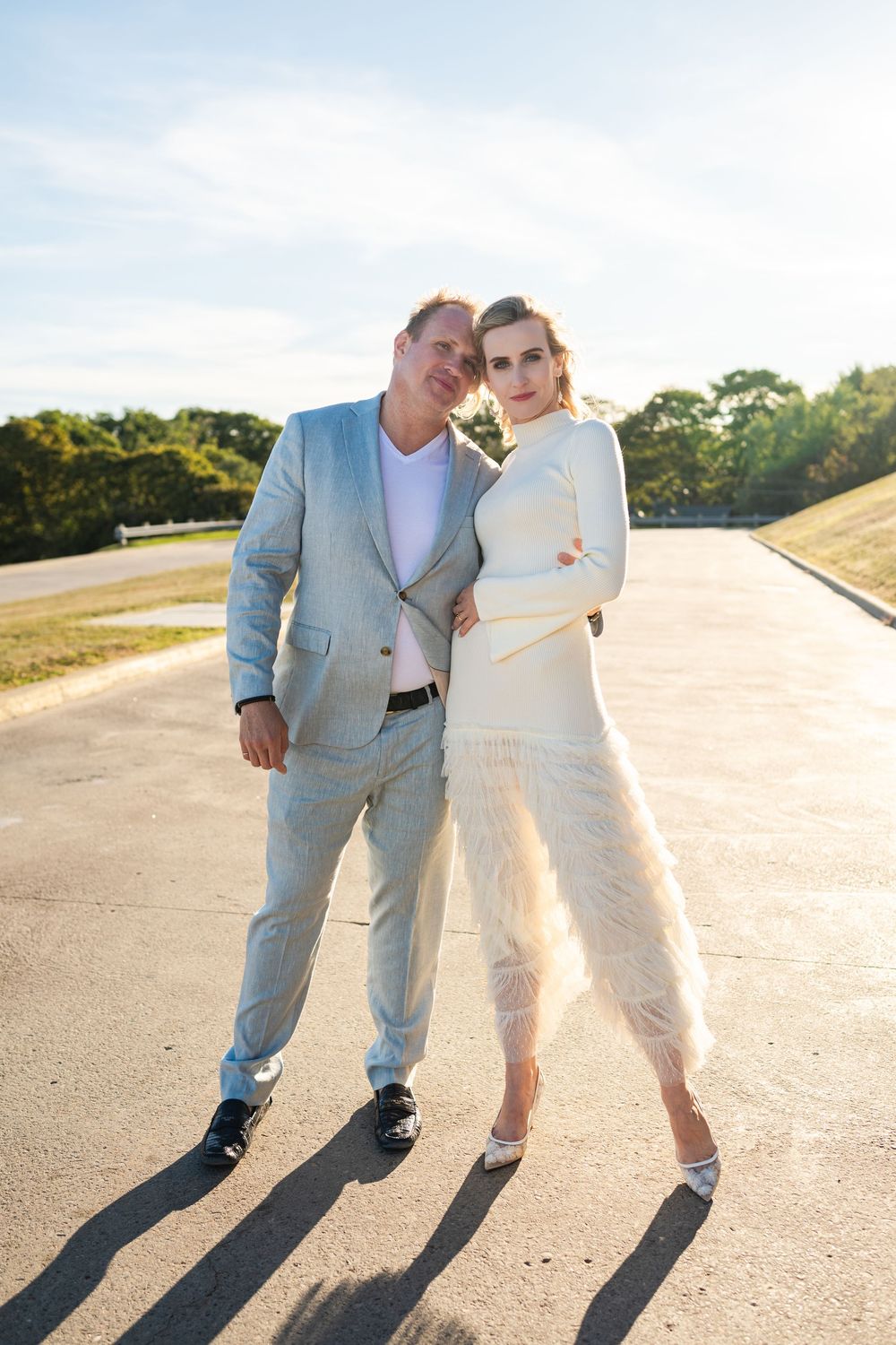Elegant couple posing outdoors on a sunny day, man in light gray suit, woman in white feathered dress.