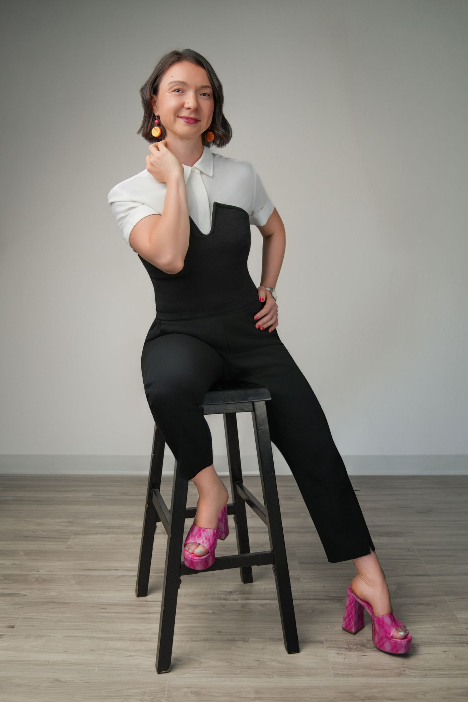 Woman in black jumpsuit and pink heels sits on stool, wearing orange earrings, posing confidently.