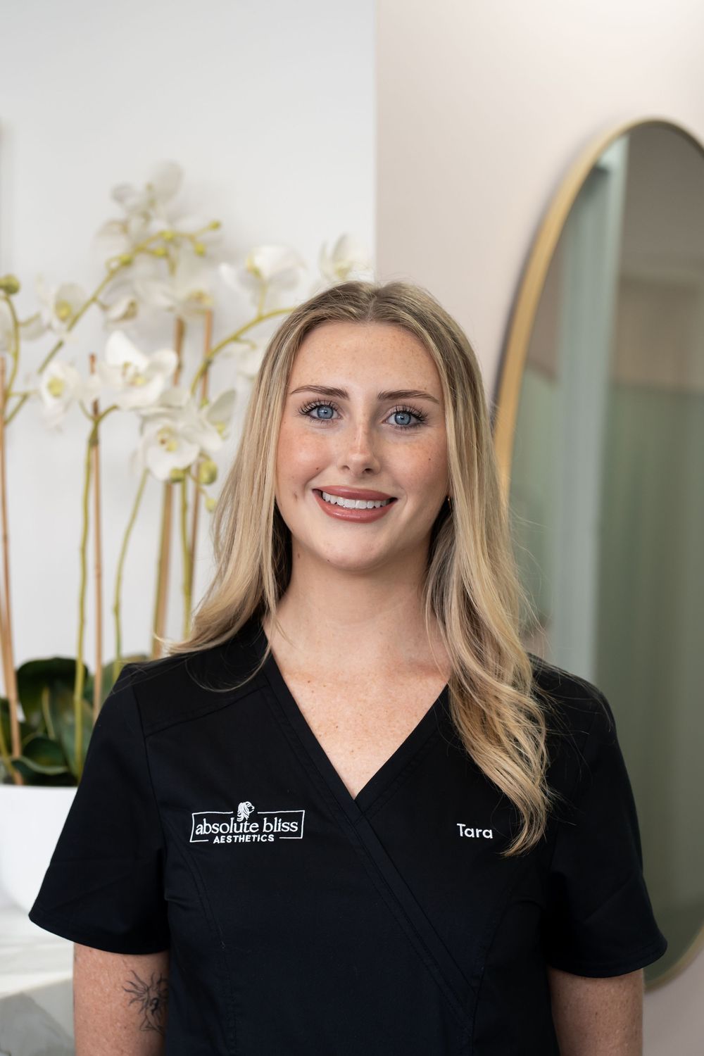 Tara, an aesthetics professional at Absolute Bliss Aesthetics, smiling in black scrubs with white orchids in background.