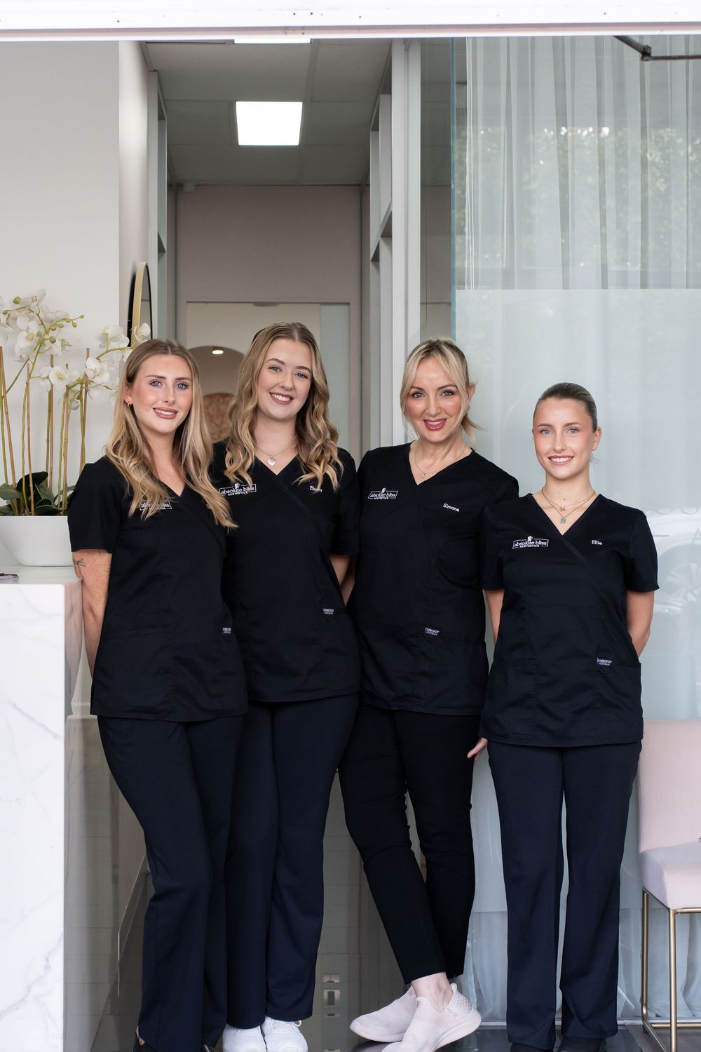Four female healthcare professionals in matching black scrubs smiling together in a modern medical spa reception area.
