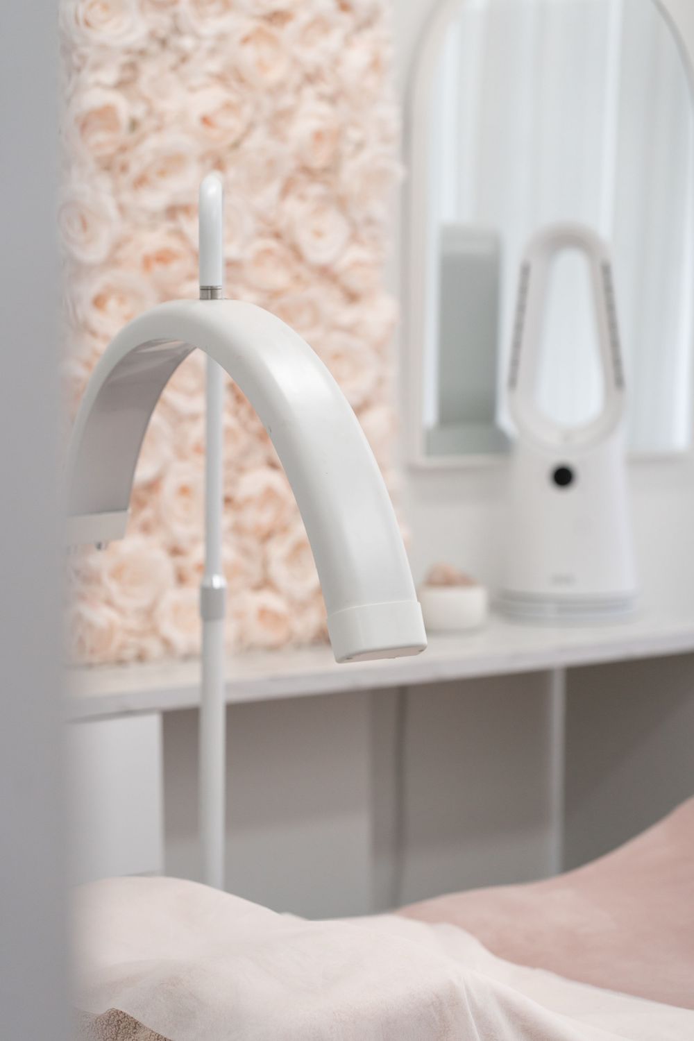 White curved beauty lamp in a modern spa room with floral wall backdrop and Dyson fan on shelf.