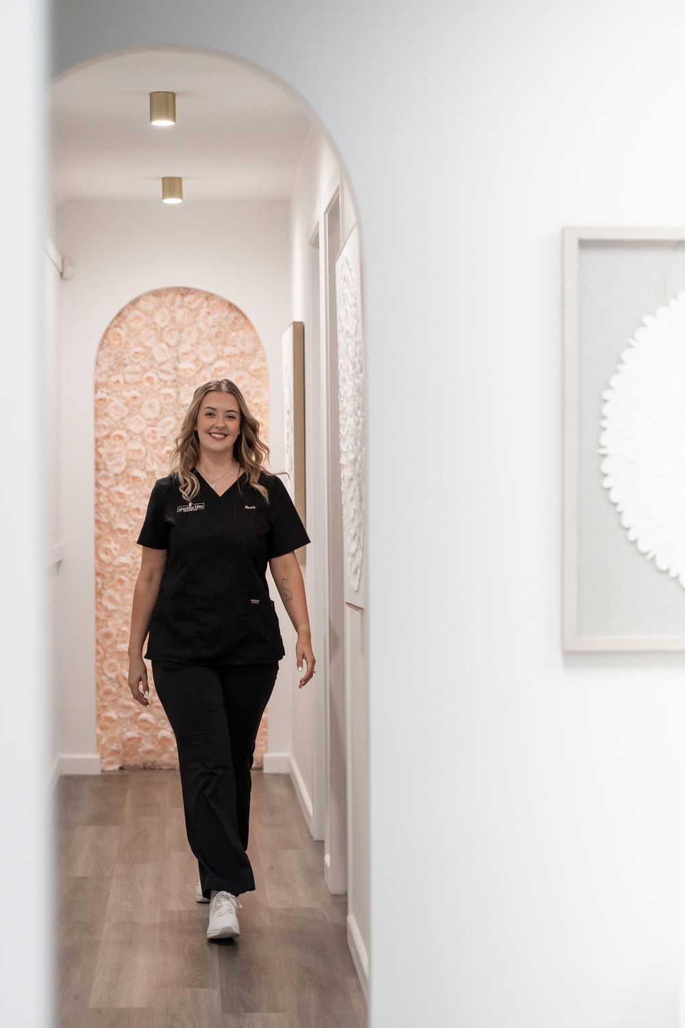 A smiling aesthetician in black scrubs walks through an elegant white arched hallway in a modern beauty clinic.