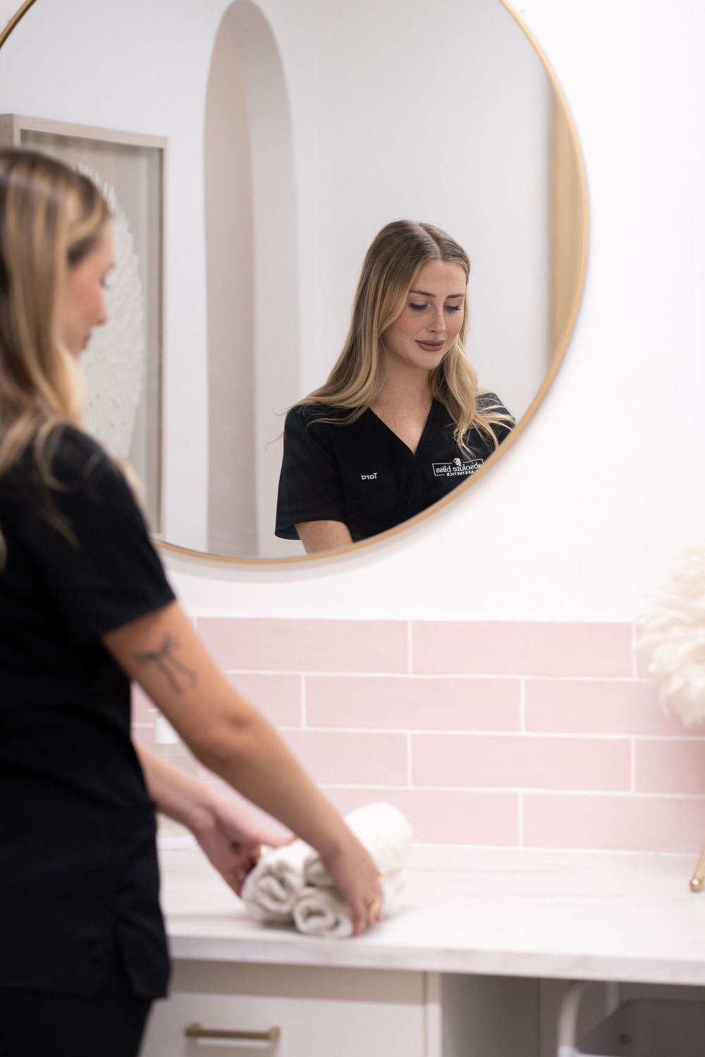 A spa professional in black scrubs arranges towels on a white vanity with pink tile backsplash, reflected in a round gold mirror.