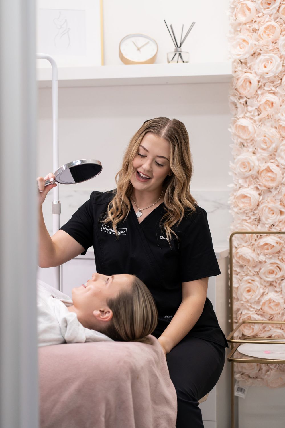 Esthetician in black scrubs smiles while showing client results with a magnifying lamp in a pink floral spa room.