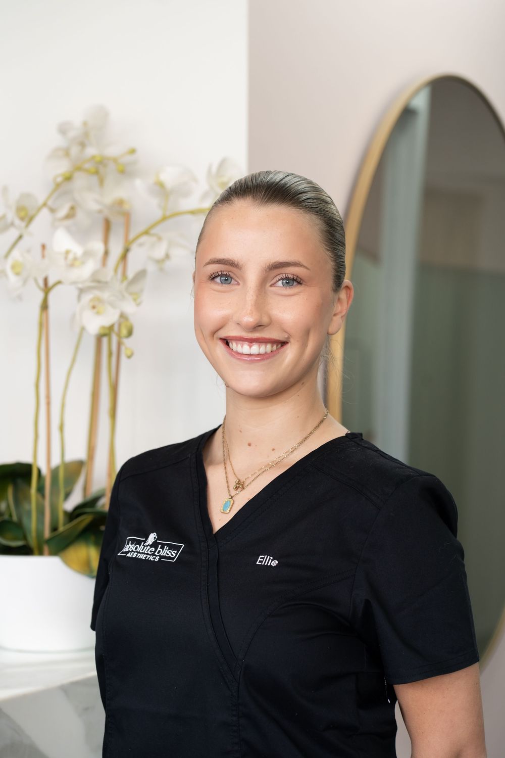 Smiling female aesthetician named Ellie in black scrubs at Jackin Bliss Aesthetics clinic with orchids.