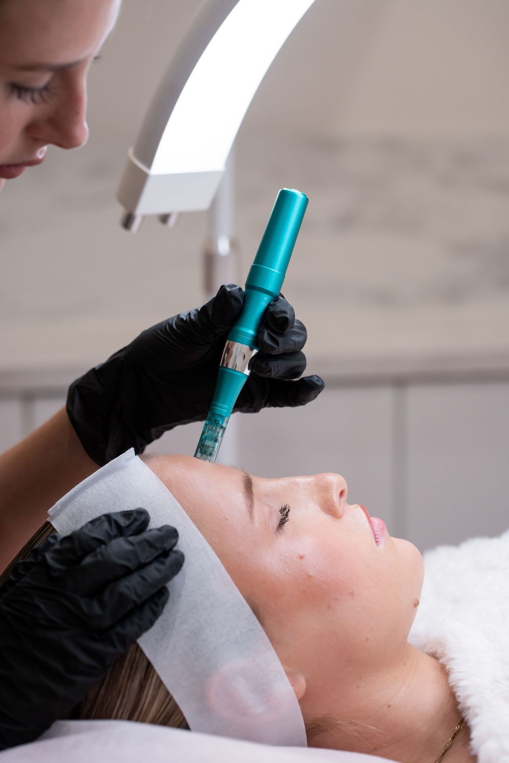 Esthetician in black gloves performing microneedling treatment on a female client using a teal dermapen device.