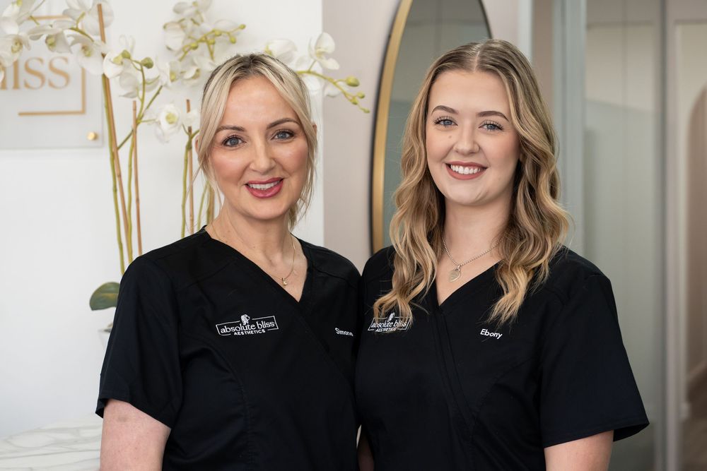 Two smiling women in black medical scrubs pose together in a modern clinic reception area.