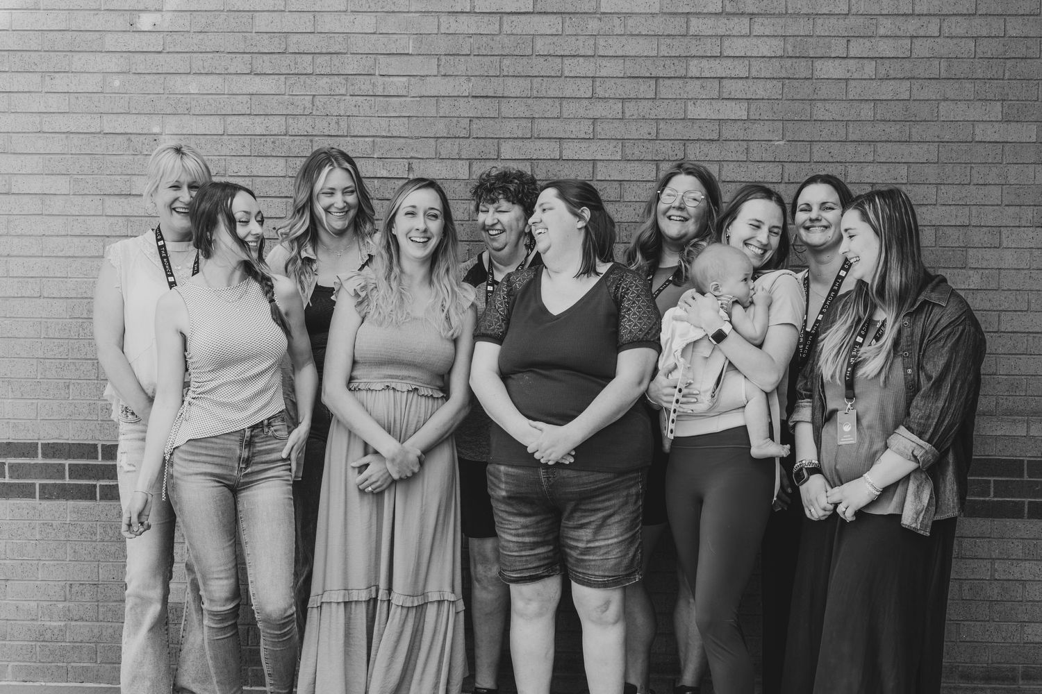 Black and white photo of a group of nine women smiling and laughing together against a brick wall.