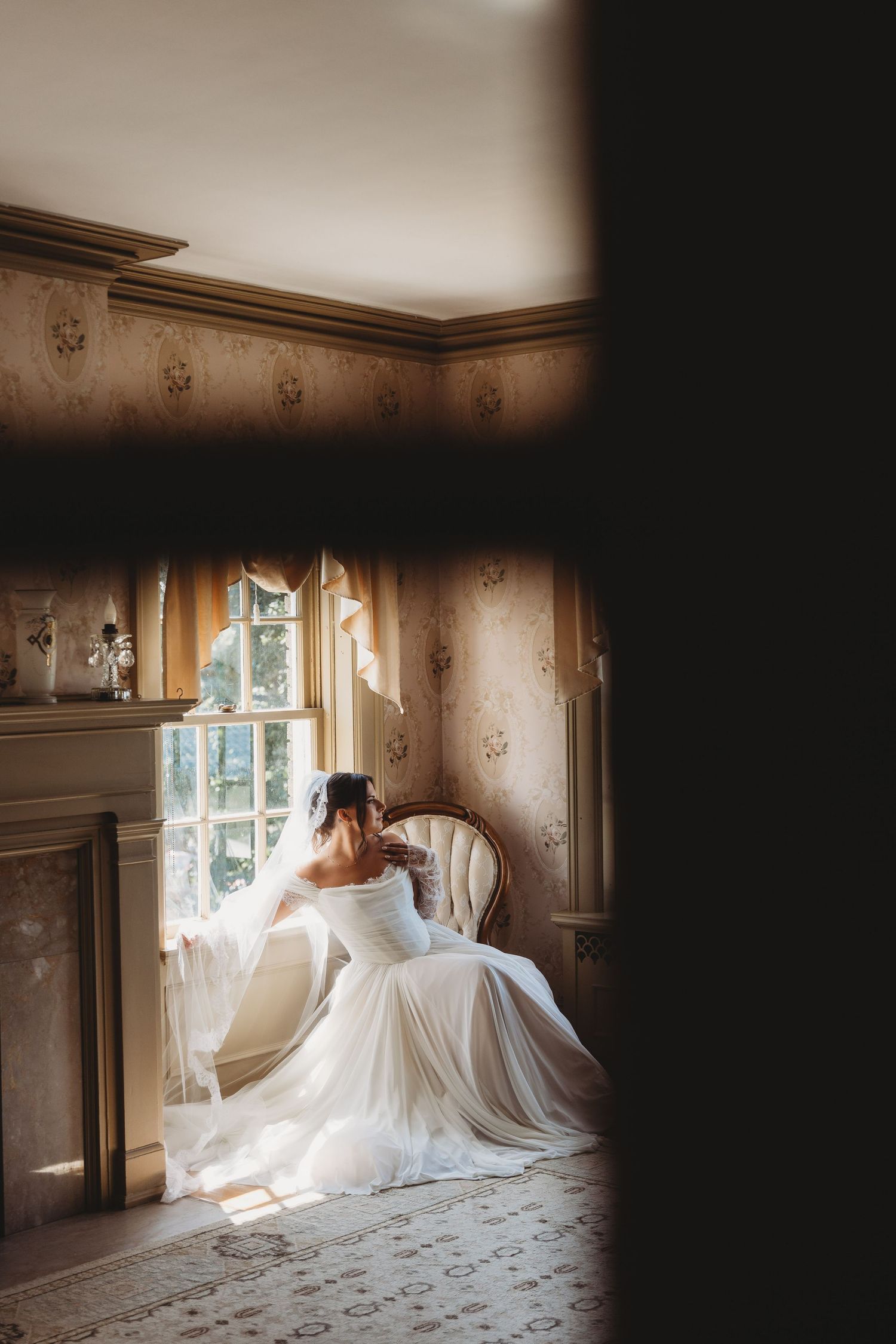 Bride in white gown sits by sunlit window in elegant vintage room with floral wallpaper, viewed through doorway.