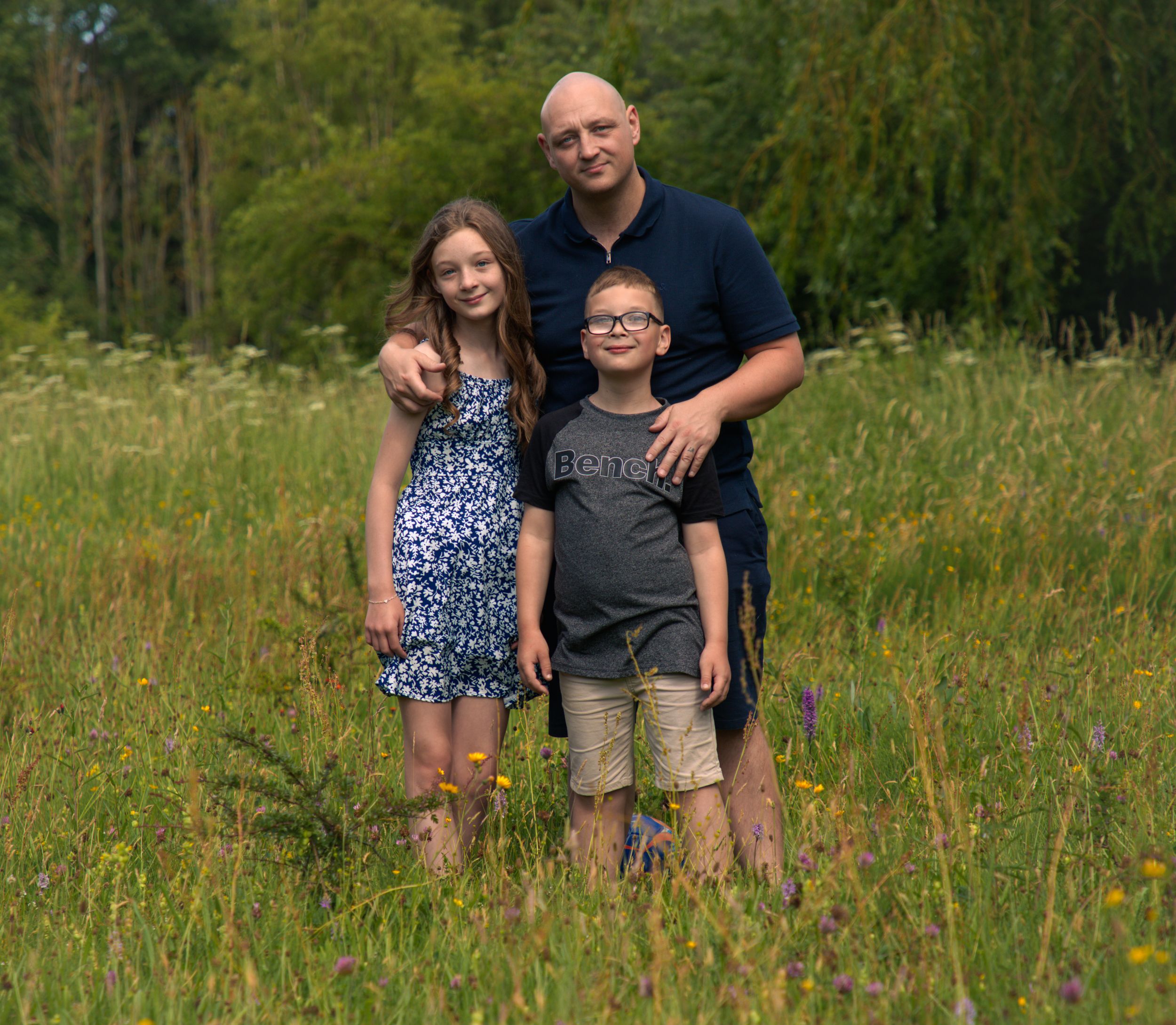 A father poses with his two children in a lush green wildflower meadow during a family portrait session.