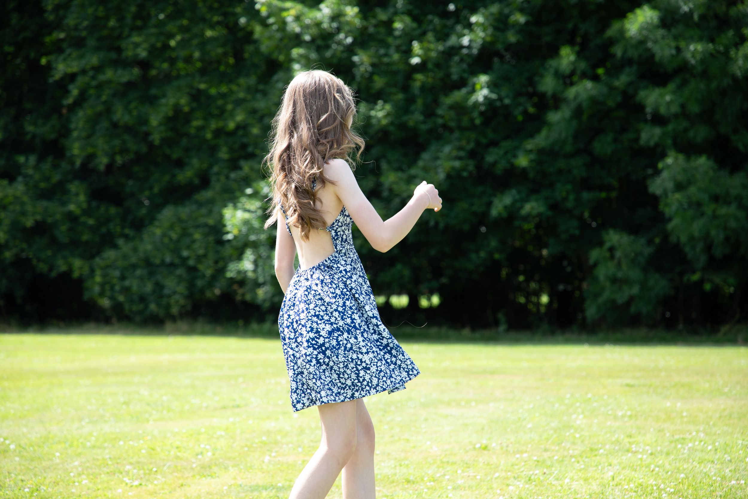 A young girl in a blue floral sundress twirls in a sunny green park surrounded by trees.