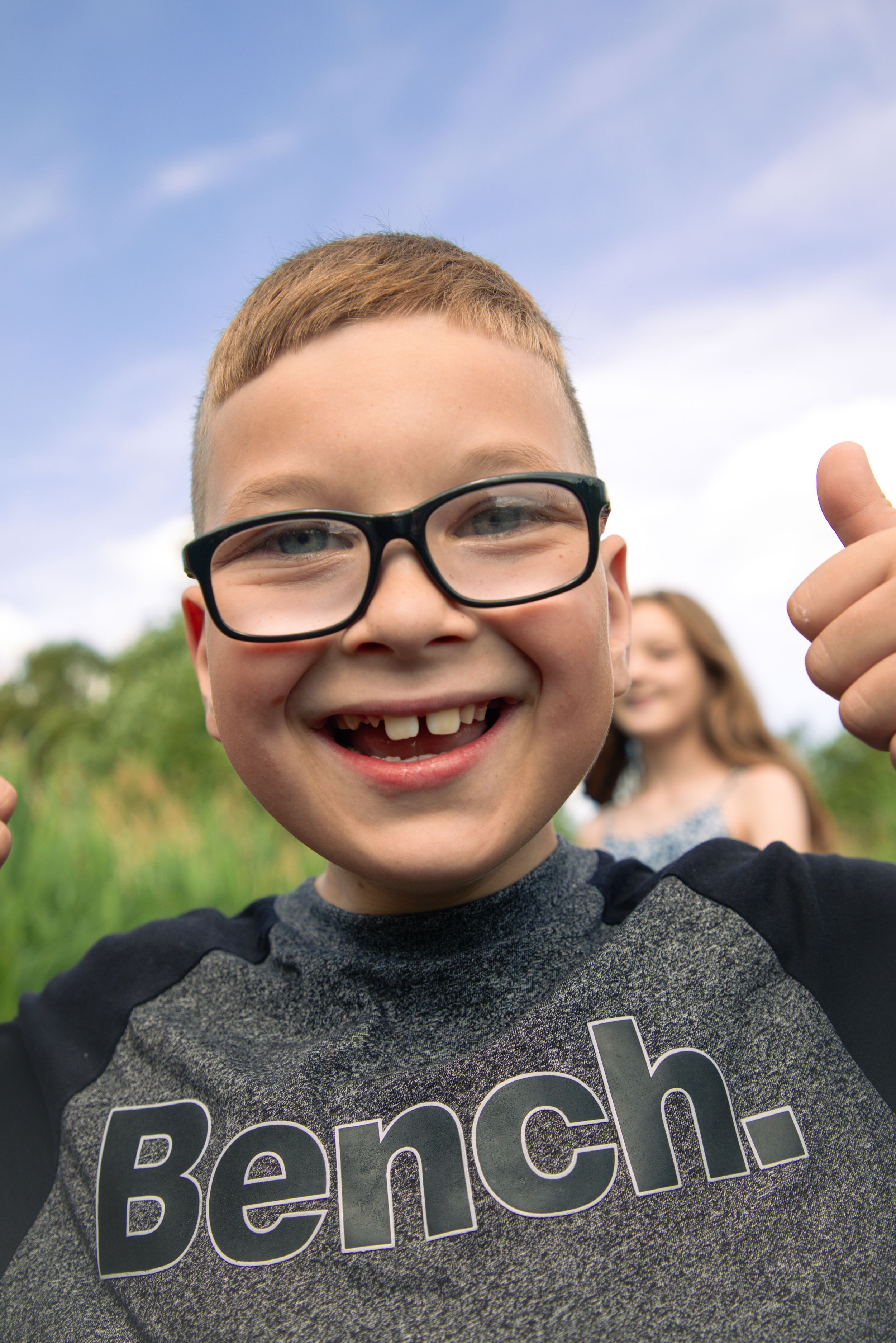 Happy young boy with black glasses giving a thumbs up wearing a gray Bench t-shirt outdoors on a sunny day.