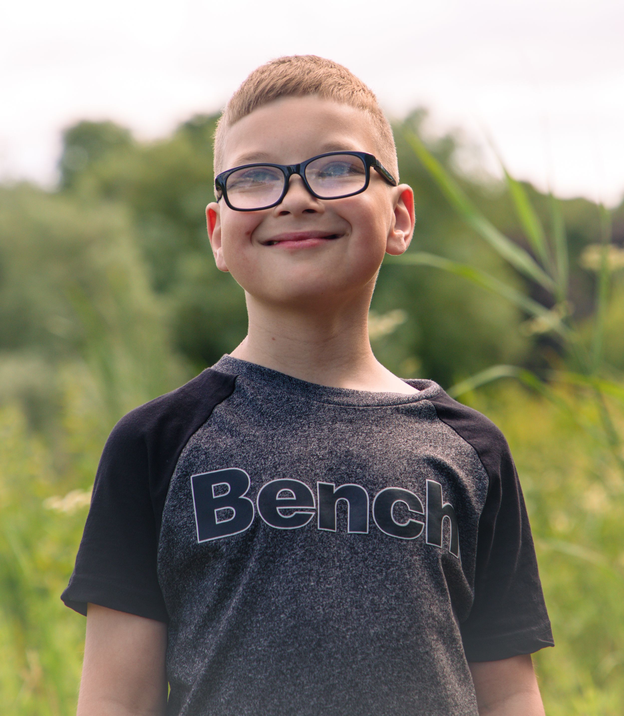Smiling boy wearing black glasses and a gray Bench t-shirt standing outdoors with green foliage in the background.