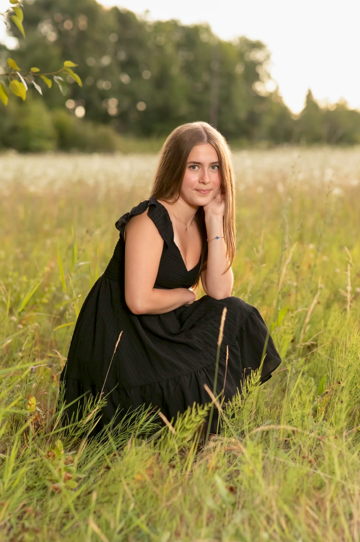 Young woman in black dress sitting gracefully in a sunlit meadow with lush green trees in the background.