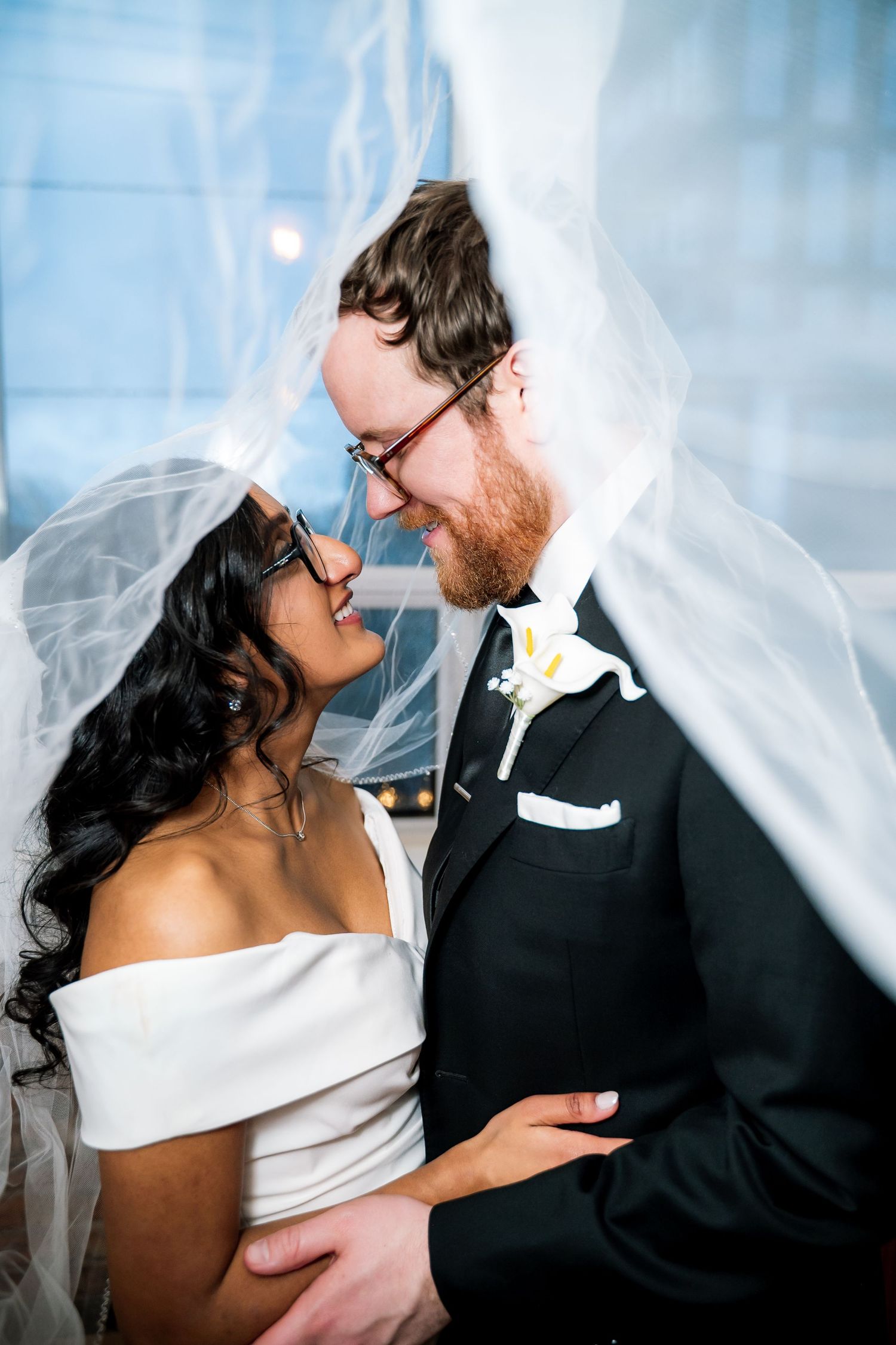 Bride and groom share a romantic moment under a flowing veil, smiling at each other in wedding attire.