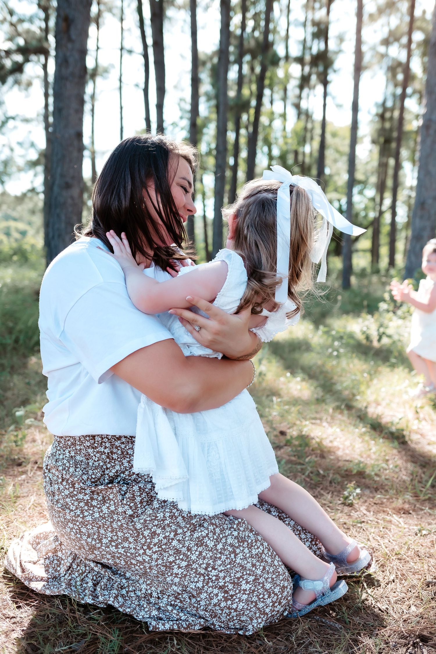 A mother kneeling in a pine forest, lovingly embracing her young daughter in matching white outfits.