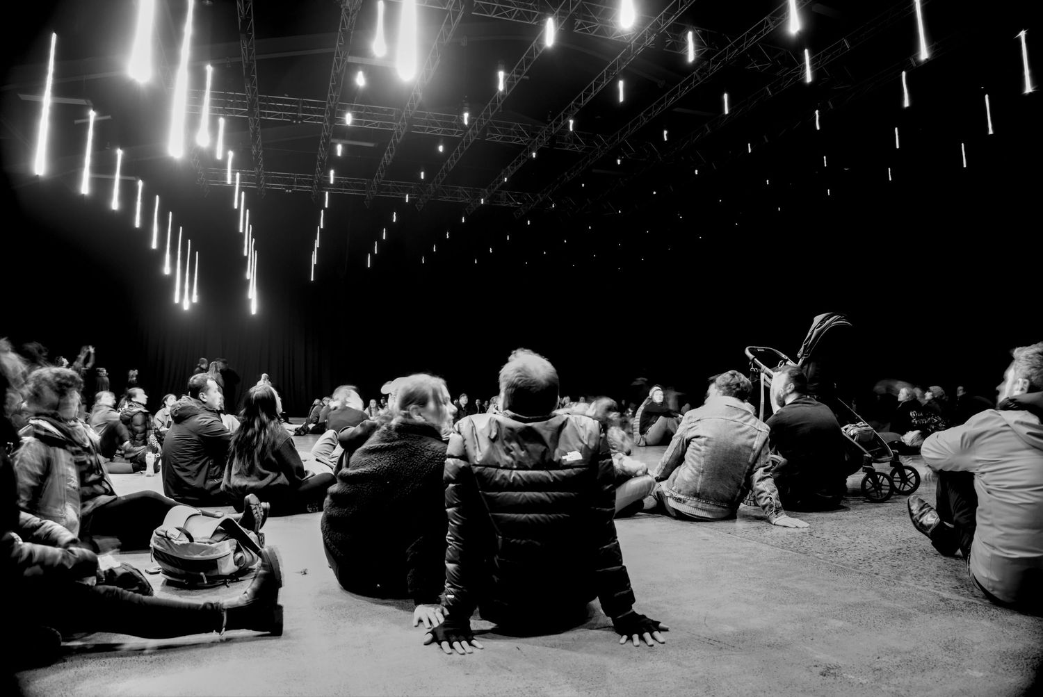 Black and white photo of audience members sitting on floor at an indoor event with neon lights hanging from ceiling.