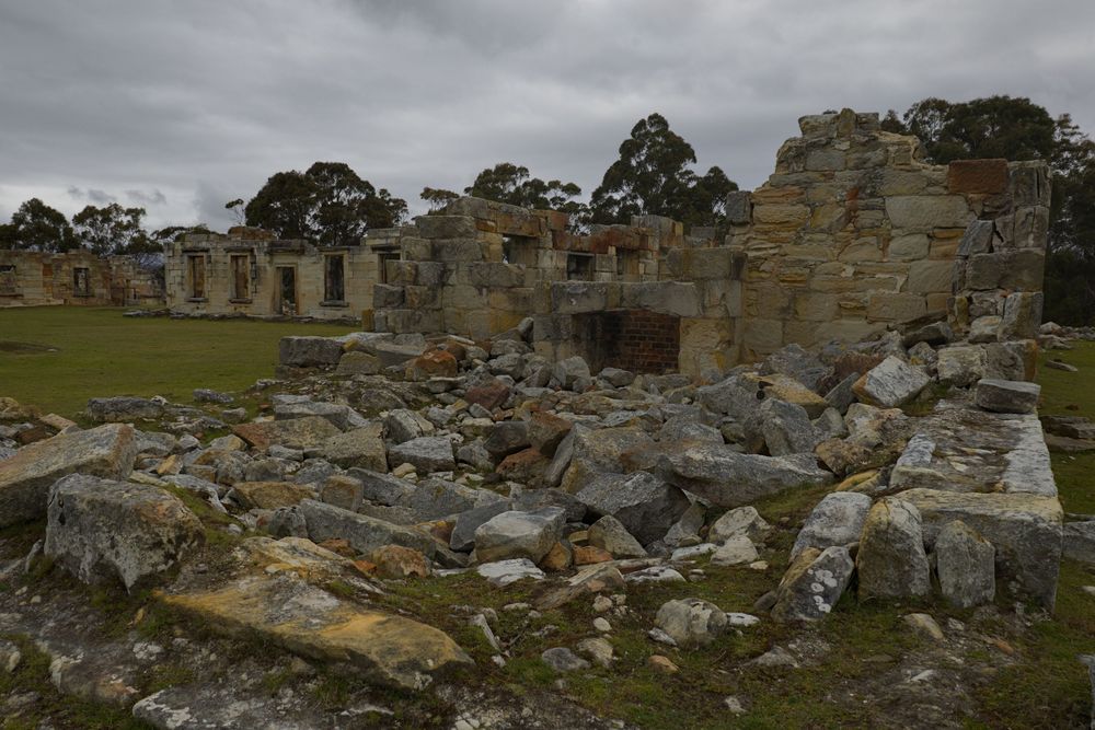 Extensive sandstone building ruins with scattered large rocks on green grass under a cloudy sky.